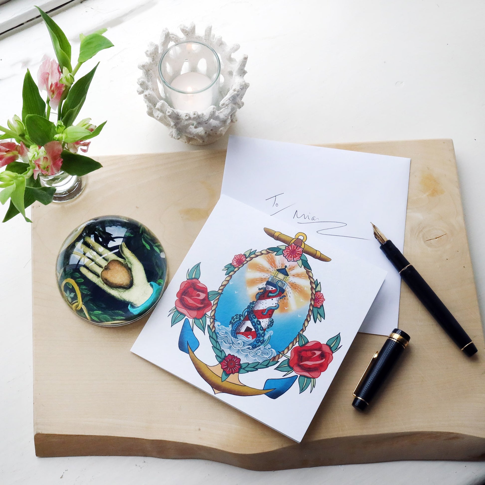 White greeting card with illustrated lighthouse & kraken tattoo inspired design on, lying on a wooden block with fountain pen, paperweight and flowers and hand written envelope.