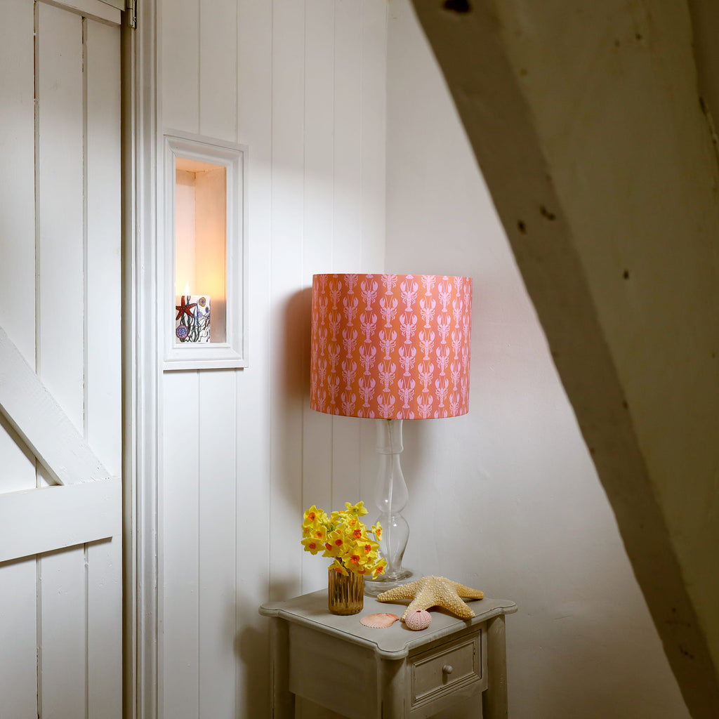 Small table with a pink patterned mini lobster lamp, flowers, and a starfish against a white wall.