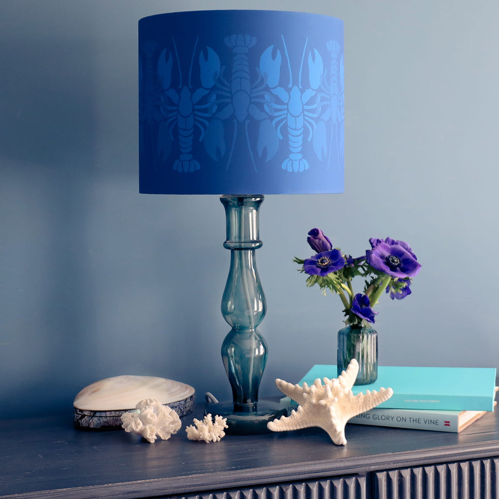 Blue lamp with lobster pattern on shade, books, and flowers on a shelf against a blue wall.