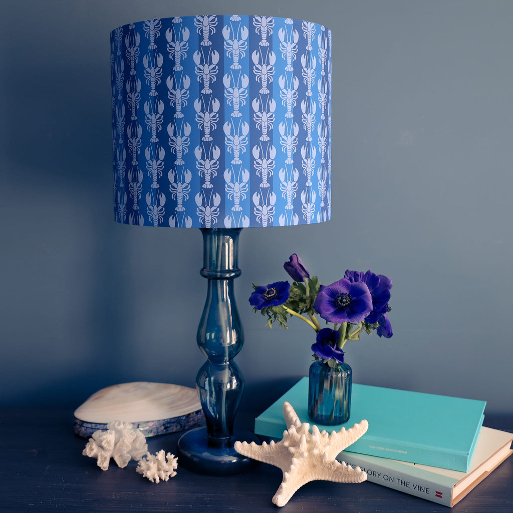 Blue lamp with lobster patterned shade on a dark surface with books and decorative items.