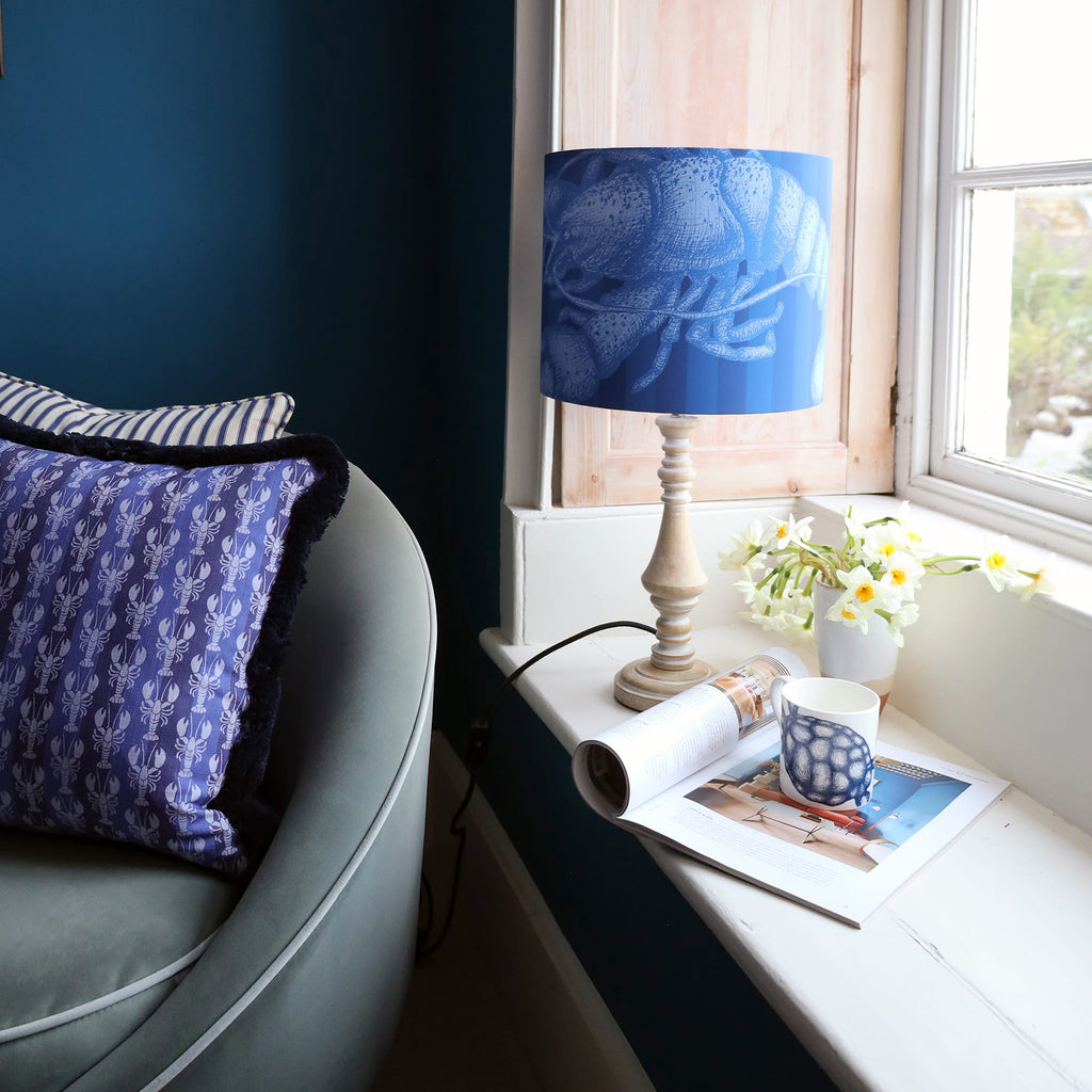 Blue armchair with a lobster-print cushion beside a window, next to a wooden lamp with a blue lobster-patterned shade, styled with a mug, flowers, and an open magazine.