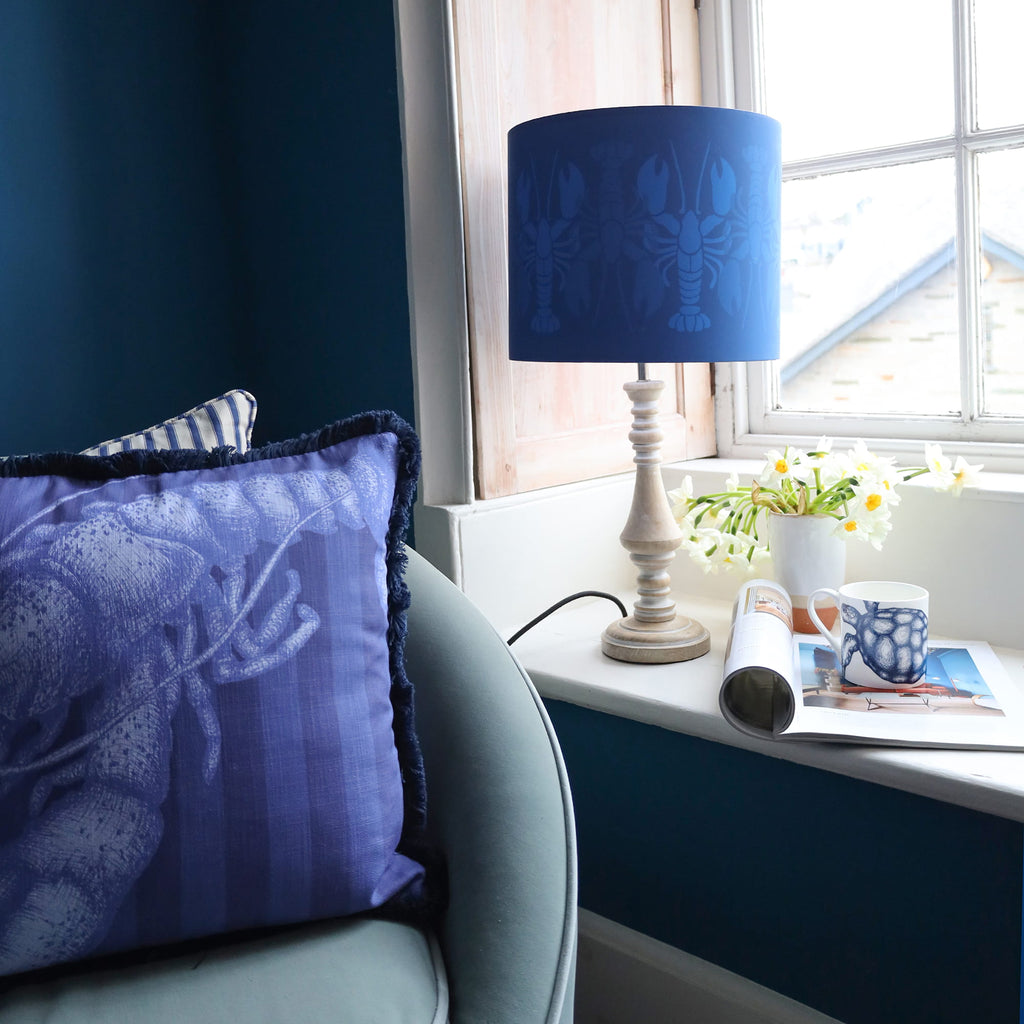 Cozy corner with a blue armchair, lobster-print cushion, and matching navy lampshade on a small table by a sunlit window, styled with flowers, a mug, and a magazine.