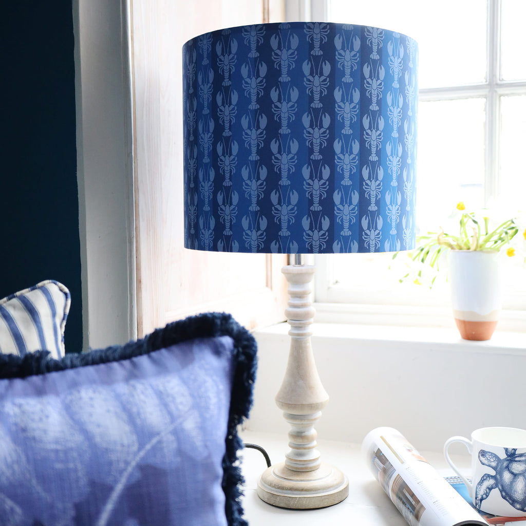 blue striped lampshade with mini lobsters on whitewash wooden turned lamp base, in window with bright sunlight behind.