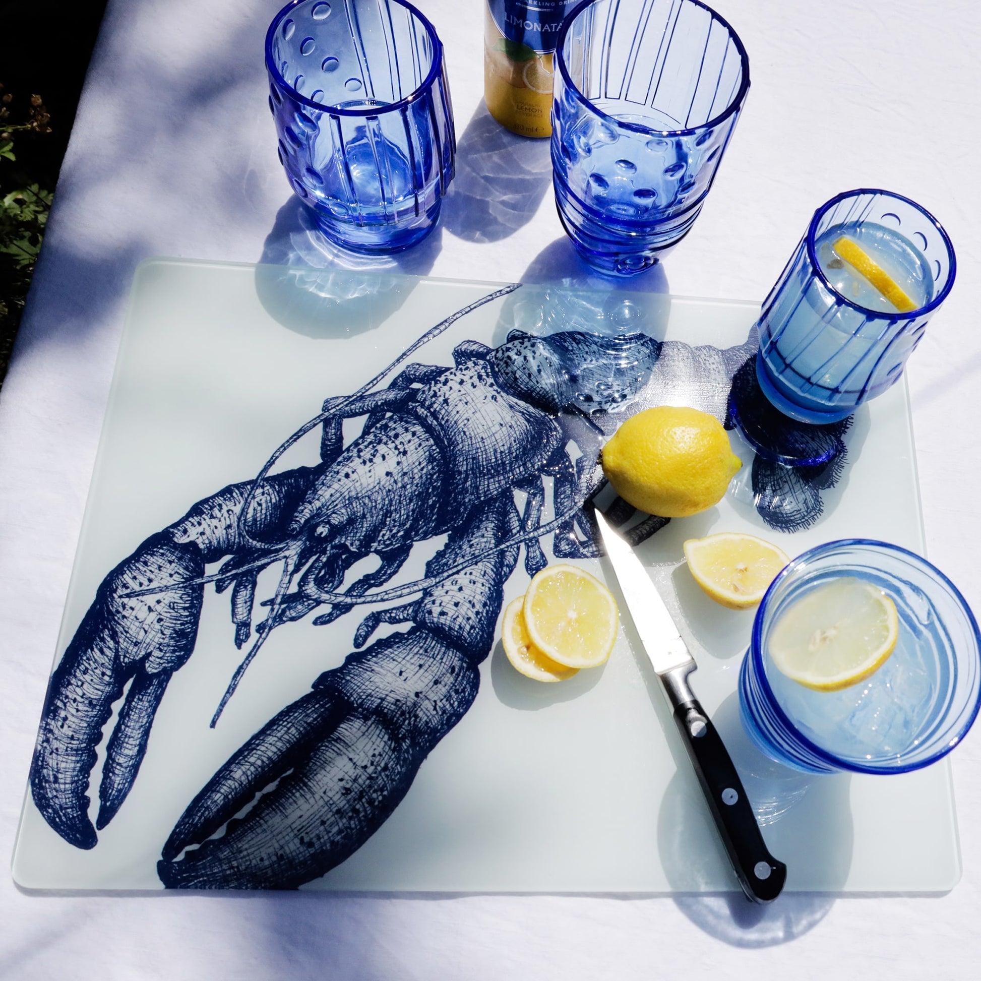 Blue-toned lobster illustration on a glass worktop saver, surrounded by blue drinking glasses, lemon slices, and a knife on a white tablecloth in bright natural light.
