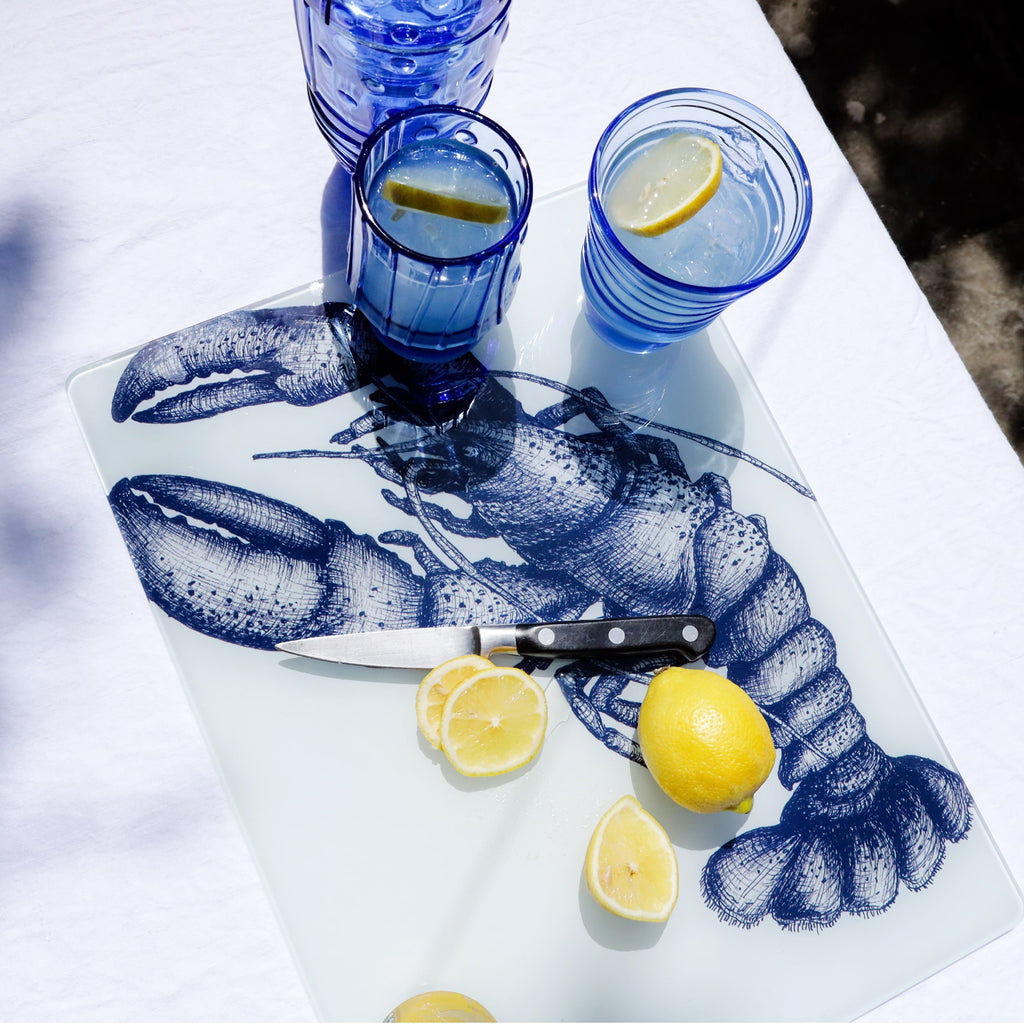 Glass worktop saver featuring a detailed blue illustration of a lobster, surrounded by blue drinking glasses with lemon slices, a kitchen knife, and fresh lemons on a white tablecloth in bright sunlight.