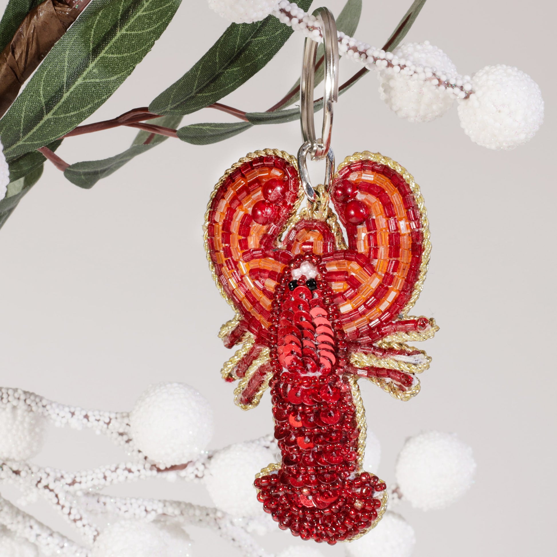 A beaded red lobster keyring with orange and gold accents hanging from a branch with green leaves and white frosted berries against a light background.