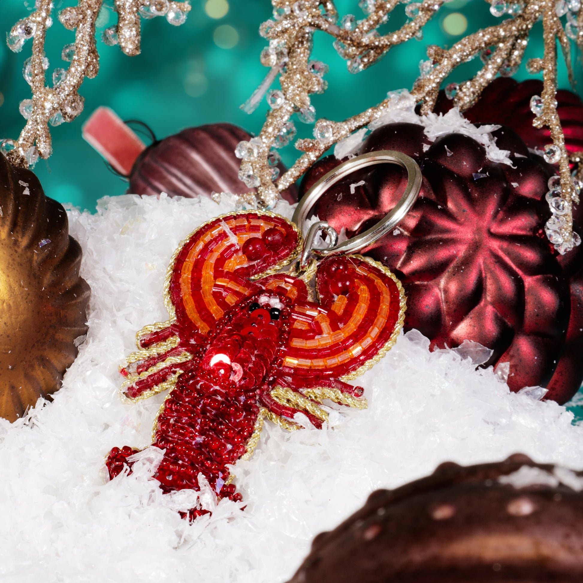 A beaded red lobster keyring with black bead eyes displayed on artificial snow among red and gold Christmas baubles and glittering gold branches against a teal background.