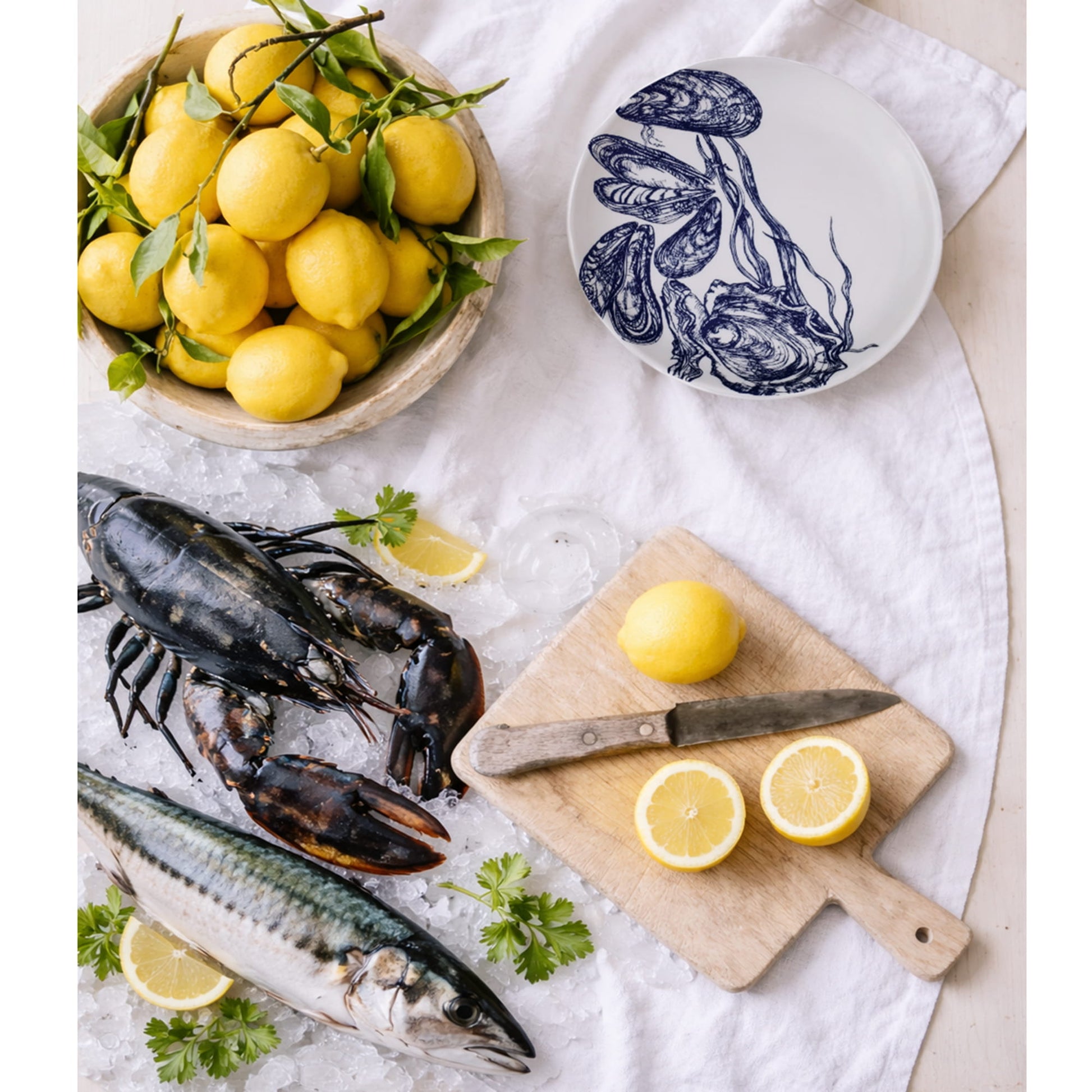 Fresh lemons, lobsters, and mackerel on a white surface with a decorative mussel & oyster plate.