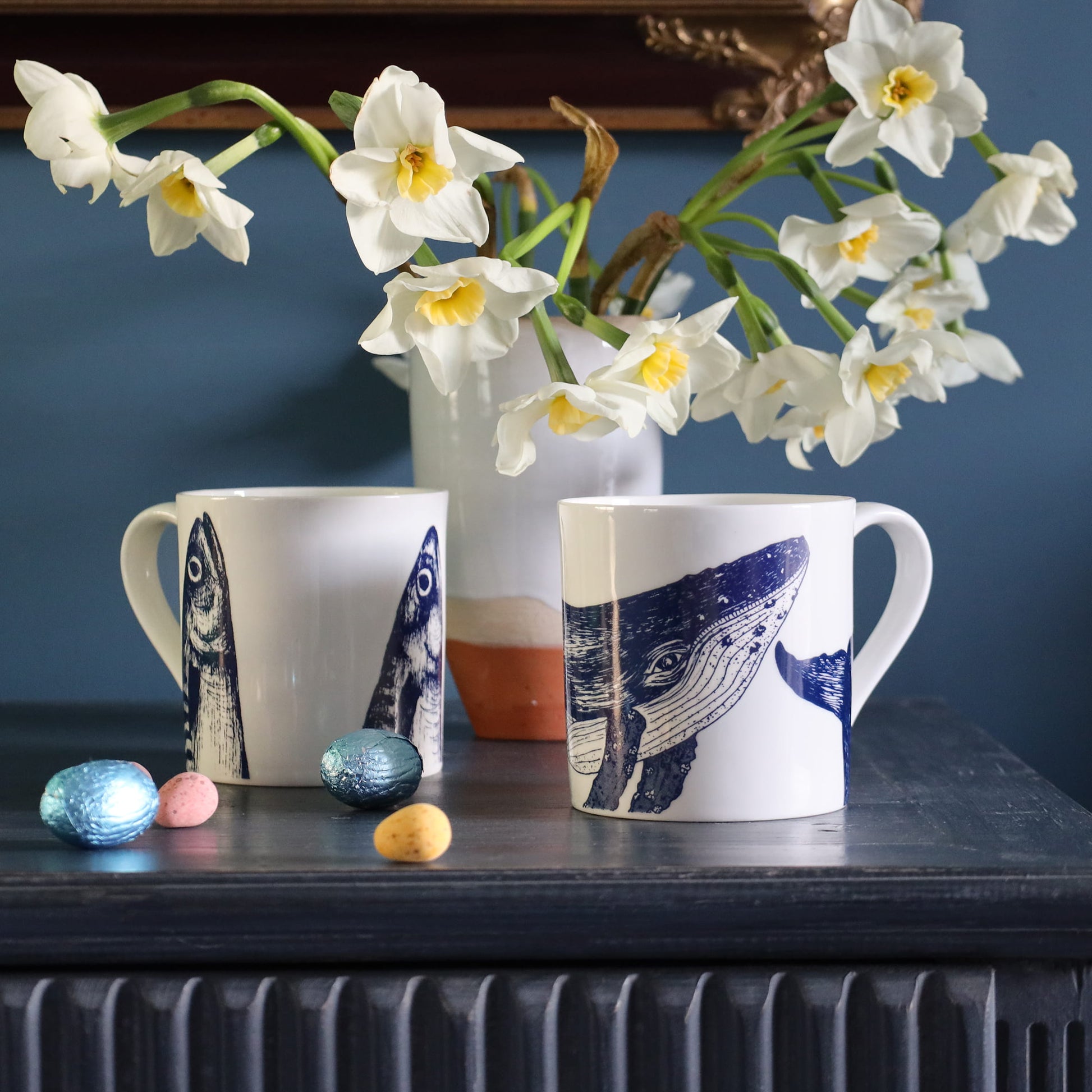 Two mugs with whale designs on a table with flowers and small objects.