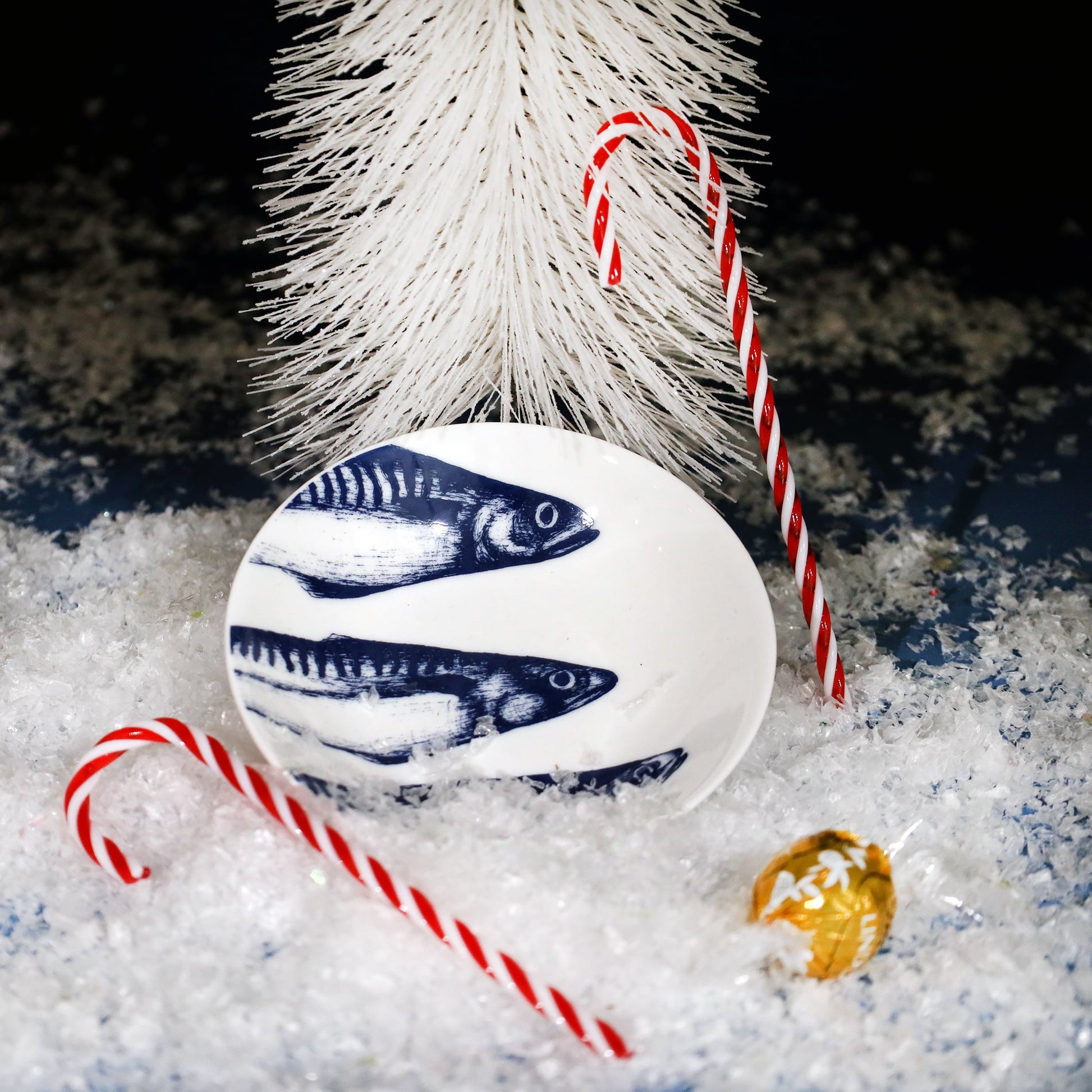 Festive scene featuring a white dish with blue mackerel illustrations, surrounded by faux snow, candy canes, and a gold-wrapped chocolate, with a white Christmas tree in the background.