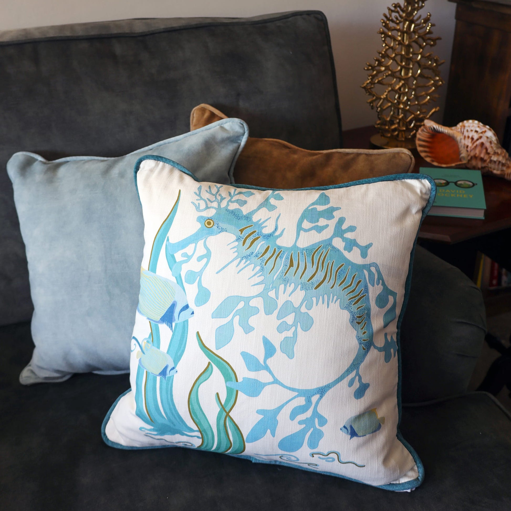 Cream Cornwall Sea Horse cushion featuring an elegant blue seahorse and coral design, styled with velvet cushions on a charcoal sofa for a relaxed coastal living room.