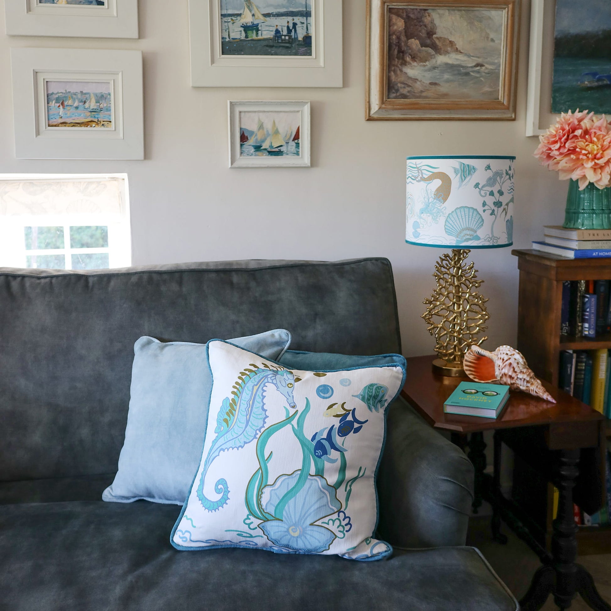 Cream Cornwall Sea Horse cushion featuring a blue seahorse, shoal of fish, seaweed and shell illustration, styled on a charcoal sofa with a velvet cushion, coastal artwork and a matching sea-inspired lampshade.
