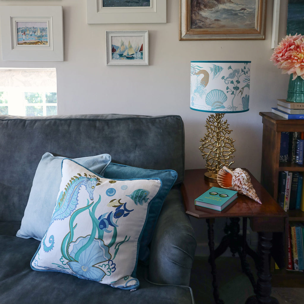 Cream Cornwall Mermaid cushion styled on a charcoal sofa with blue velvet cushions, alongside a coordinating coastal table lamp and seaside artwork for a relaxed coastal living room.