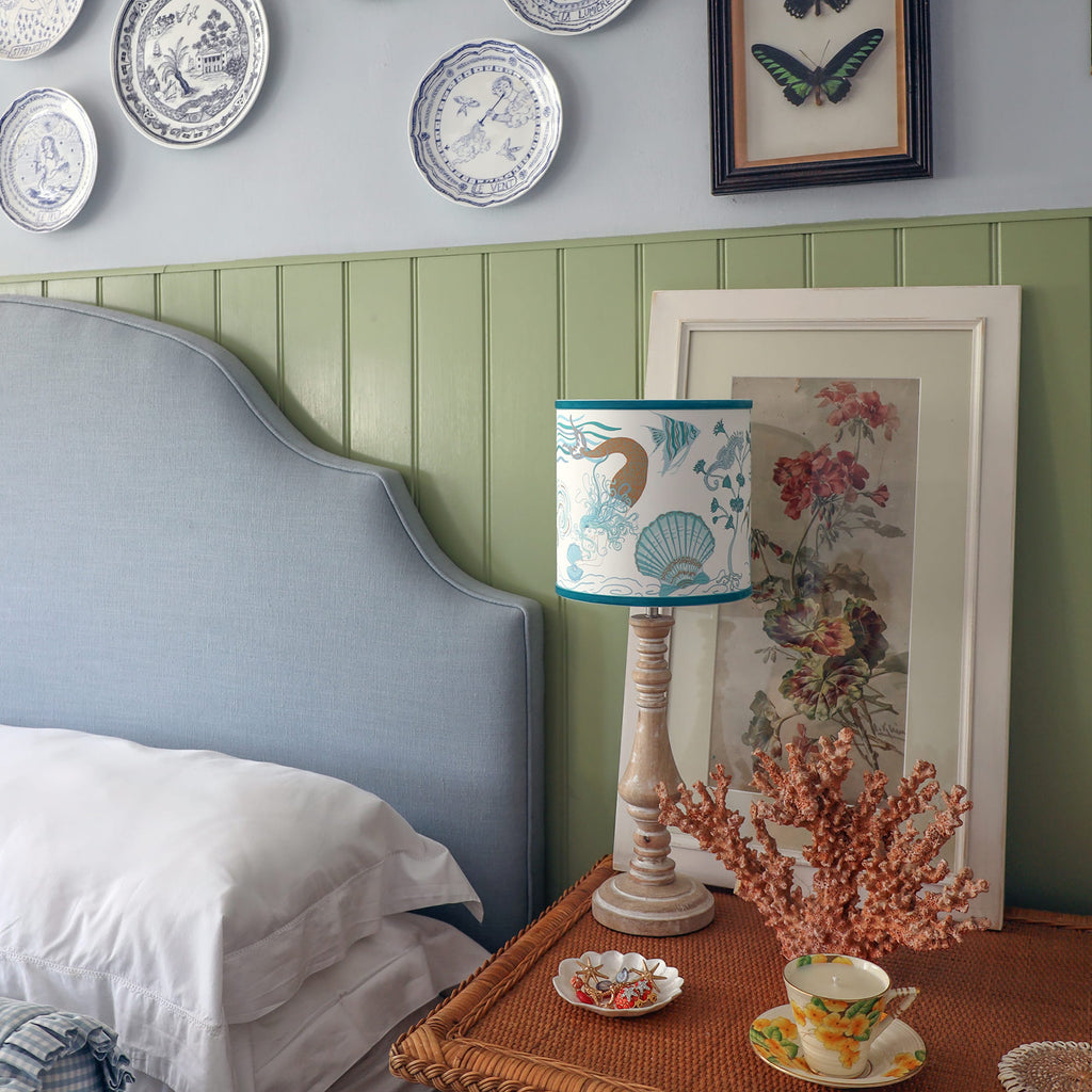 Cream Cornwall Mermaid lampshade on a turned wooden lamp base, styled on a bedside table with coral, botanical artwork and blue-and-white coastal décor in a relaxed bedroom.