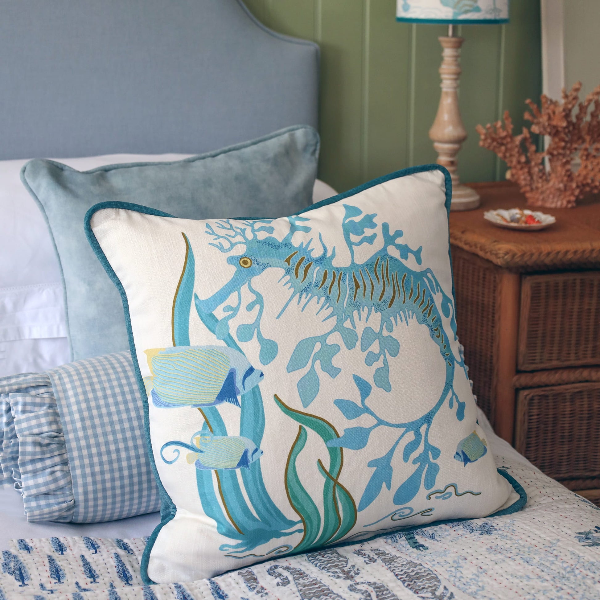 Cream Cornwall Sea Horse cushion styled on a bed with blue velvet and gingham cushions, alongside a coordinating Mermaid lampshade for a calm coastal-inspired bedroom.