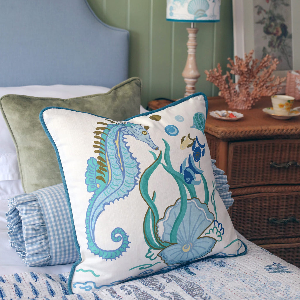 Cream Cornwall Sea Horse cushion featuring a blue seahorse, shoal of fish, seaweed and shell illustration, styled on a bed with soft velvet cushions, a gingham bolster and coastal bedroom accessories.