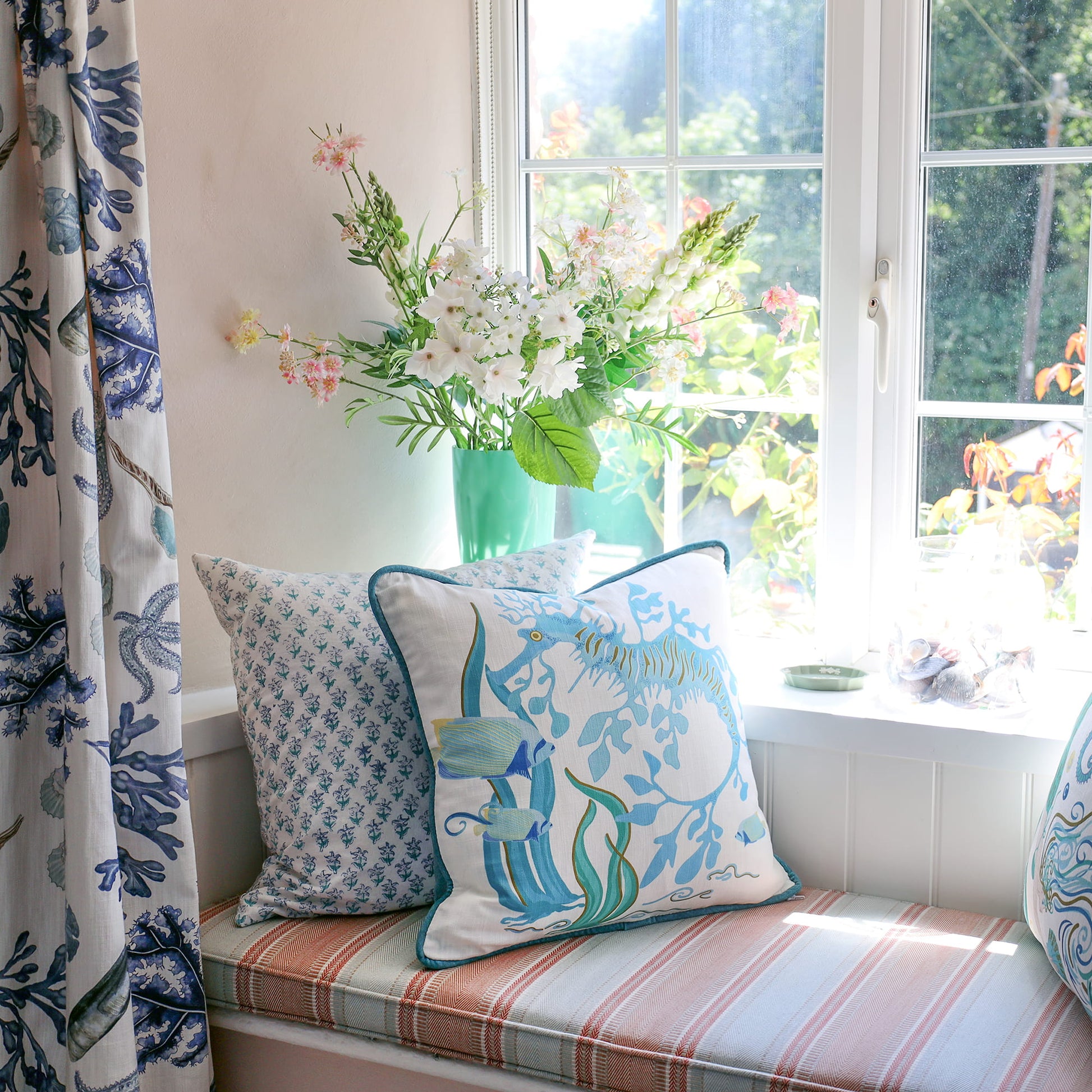 Cream Cornwall Sea Horse cushion styled on a window seat with blue patterned cushions and coordinating coastal curtains, creating a bright and relaxed seaside-inspired home.