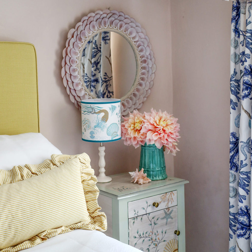 Cream Cornwall Mermaid lampshade on a bedside table, styled with a shell mirror, fresh flowers and coastal furnishings in a bright seaside-inspired bedroom.