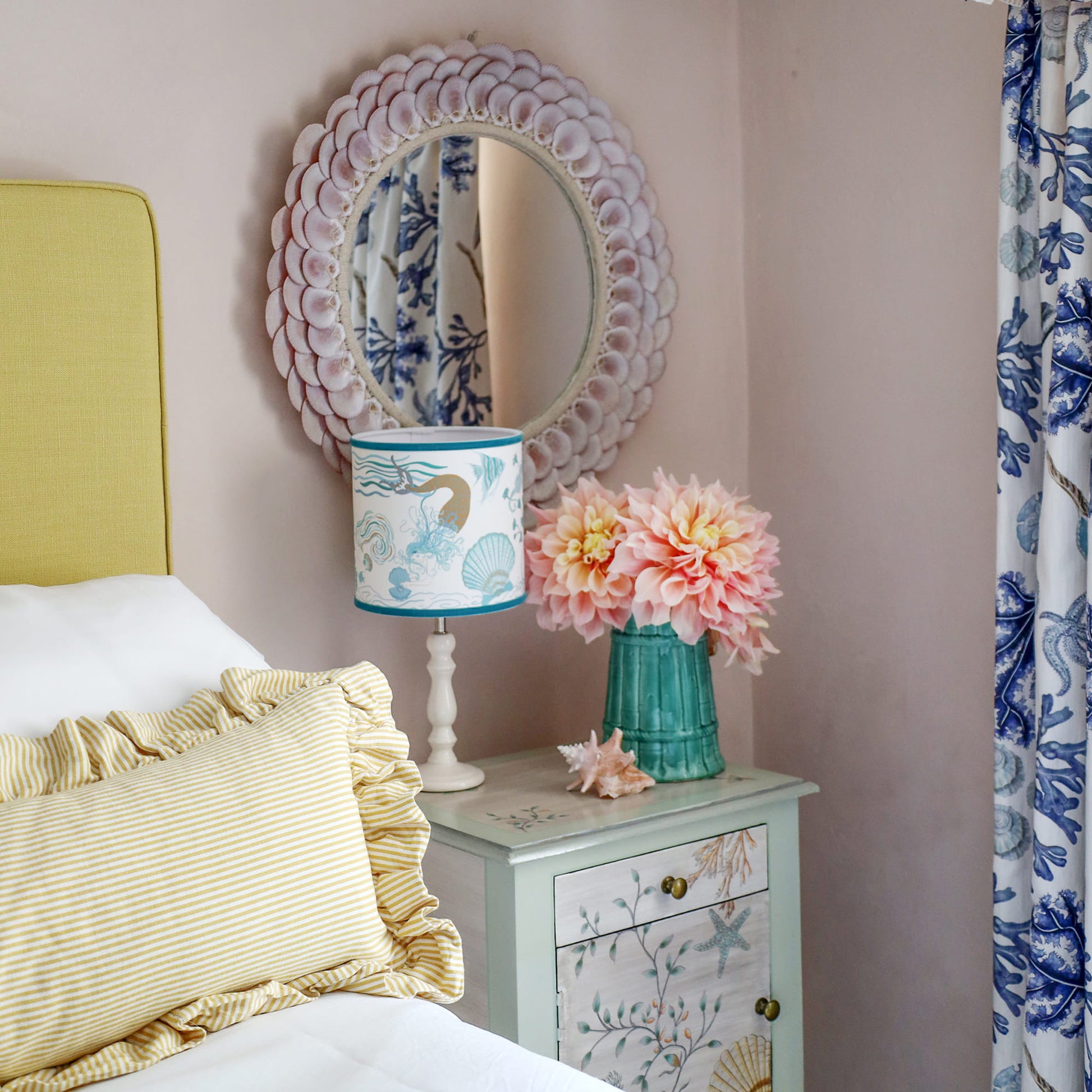 Cream Cornwall Mermaid lampshade on a bedside table, styled with a shell mirror, fresh flowers and coastal furnishings in a bright seaside-inspired bedroom.