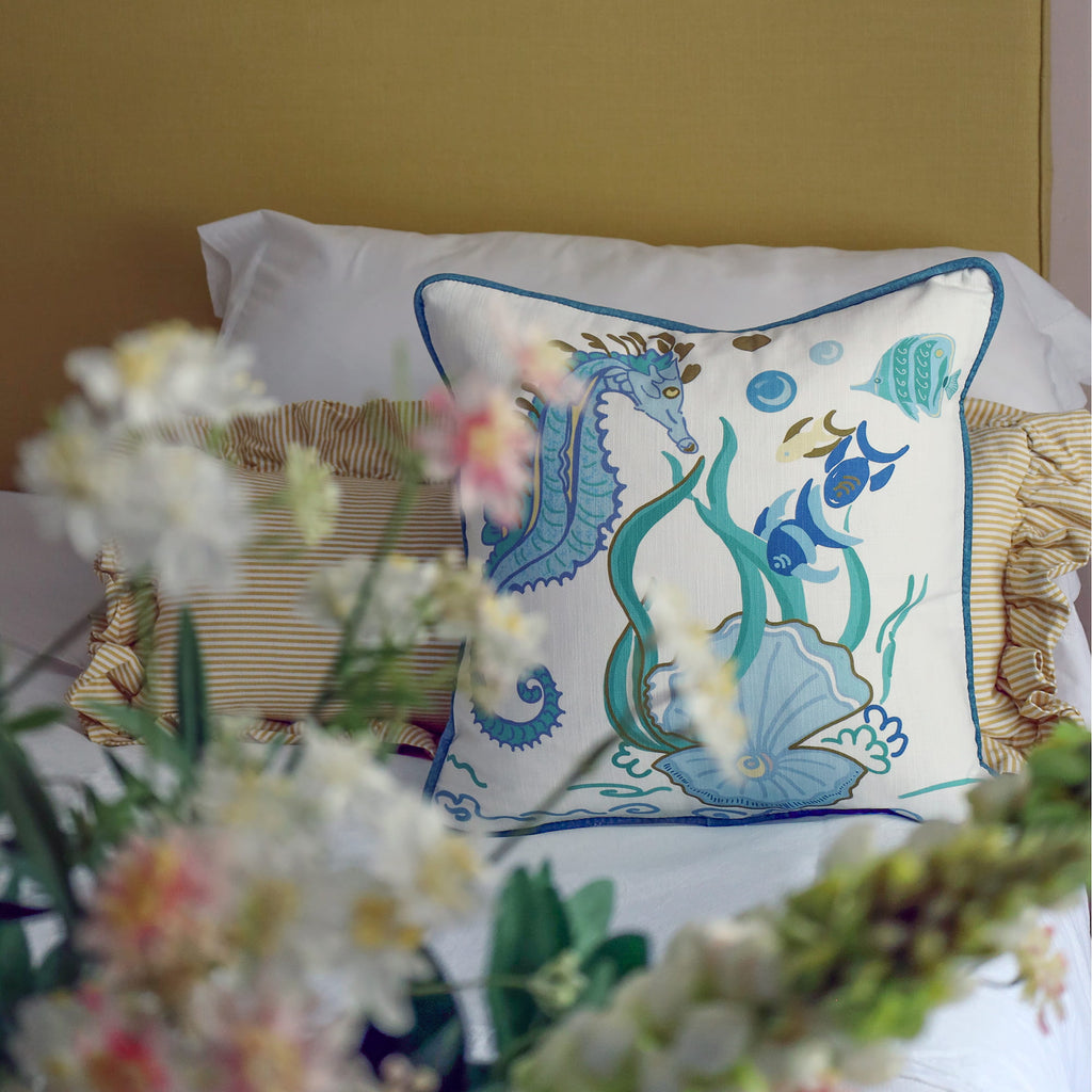 Cream Cornwall Sea Horse cushion featuring a blue seahorse, fish, seaweed and shell illustration, styled on a bed with striped ruffle cushions and soft floral accents for a fresh coastal bedroom.
