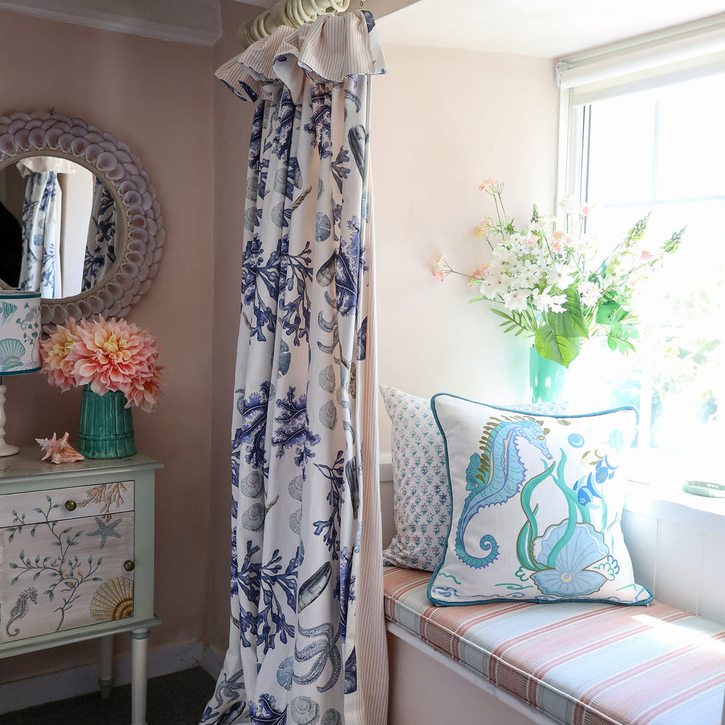 Cream Cornwall Sea Horse cushion featuring a blue seahorse, shoal of fish, seaweed and shell illustration, styled on a window seat with coastal curtains, fresh flowers and a painted bedside cabinet for a bright coastal bedroom.