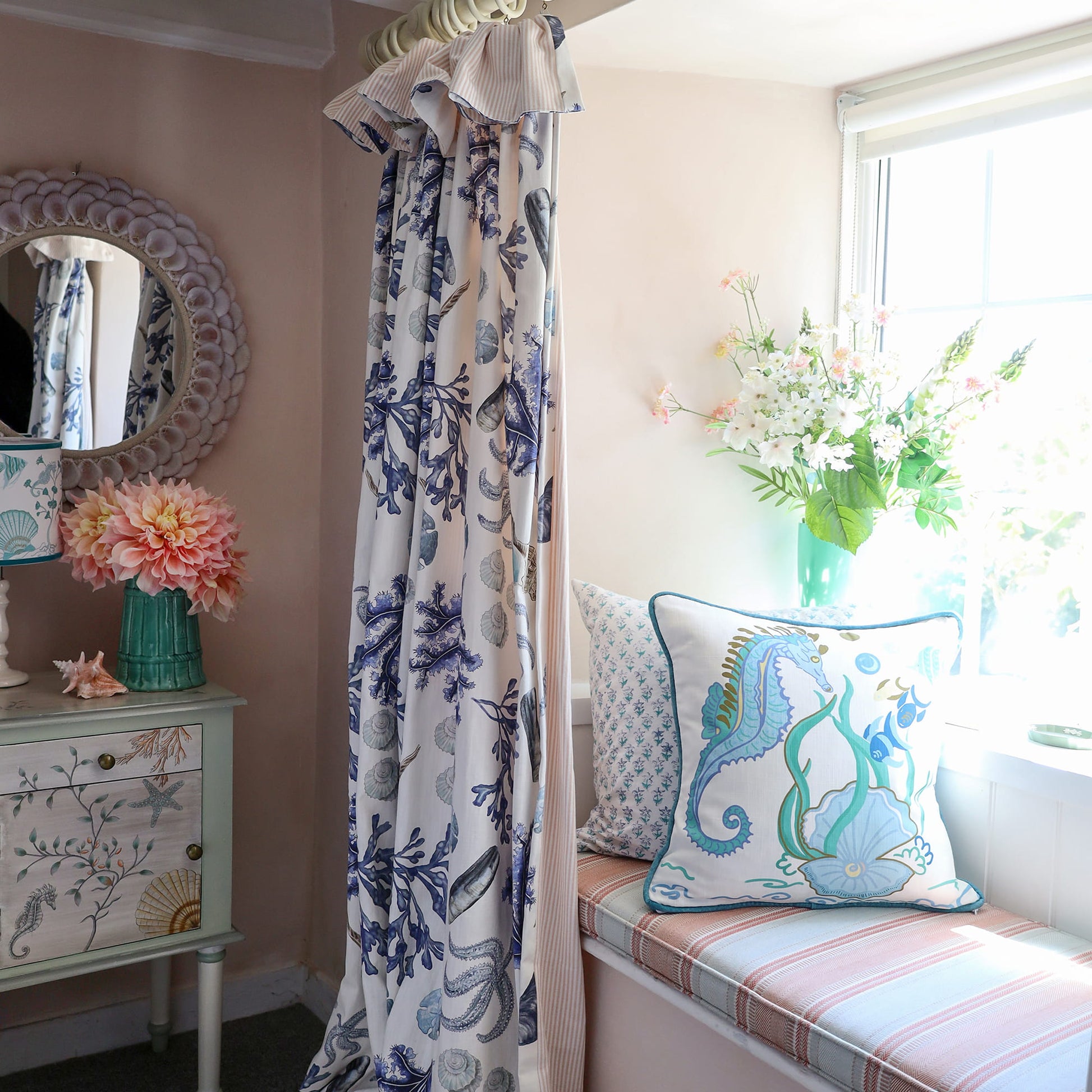 Cream Cornwall Sea Horse cushion featuring a blue seahorse, shoal of fish, seaweed and shell illustration, styled on a window seat with coastal curtains, fresh flowers and a painted bedside cabinet for a bright coastal bedroom.