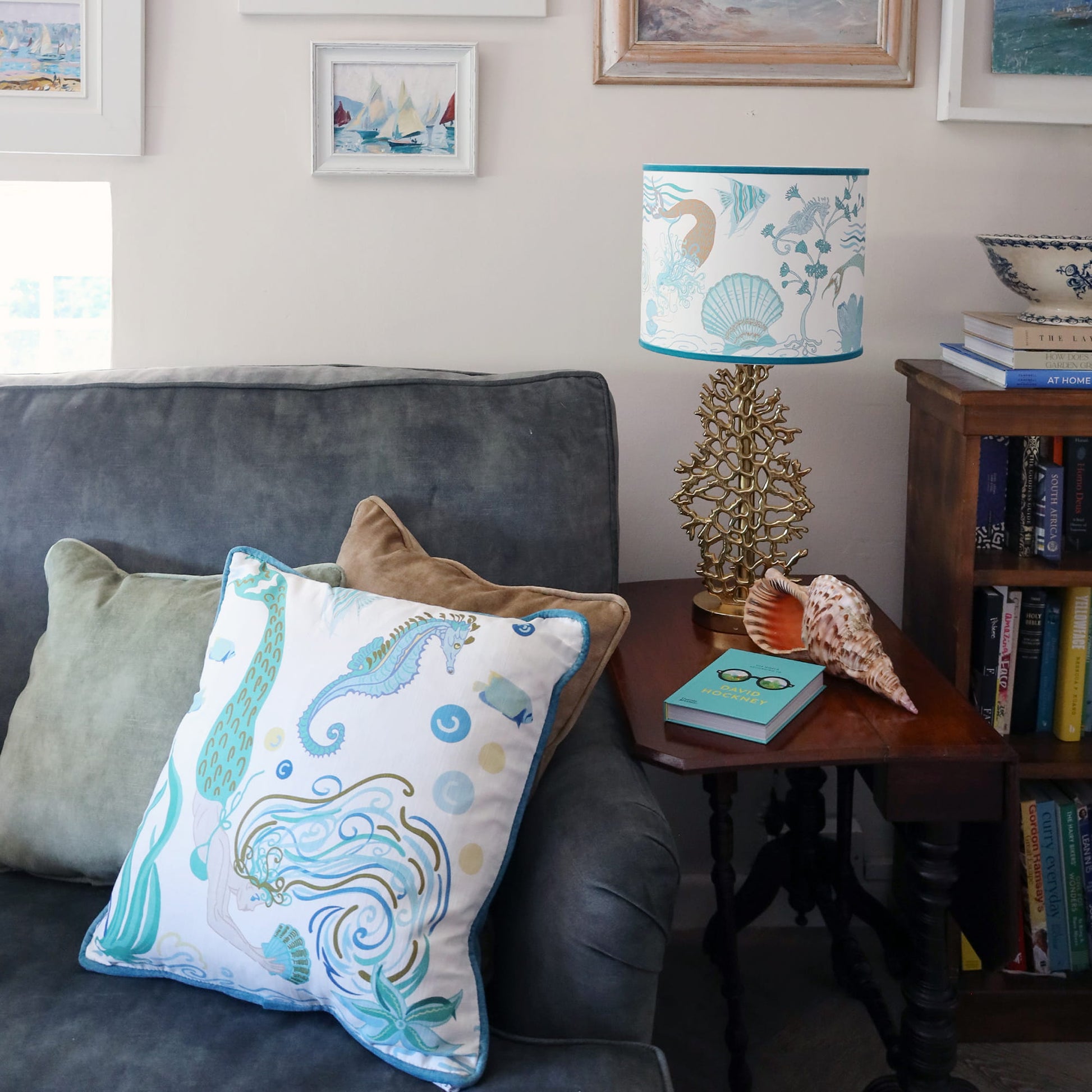 Cream Cornwall Mermaid cushion styled on a charcoal sofa with soft velvet cushions, alongside a coordinating Mermaid lampshade and coastal accessories in a relaxed seaside-inspired living room.