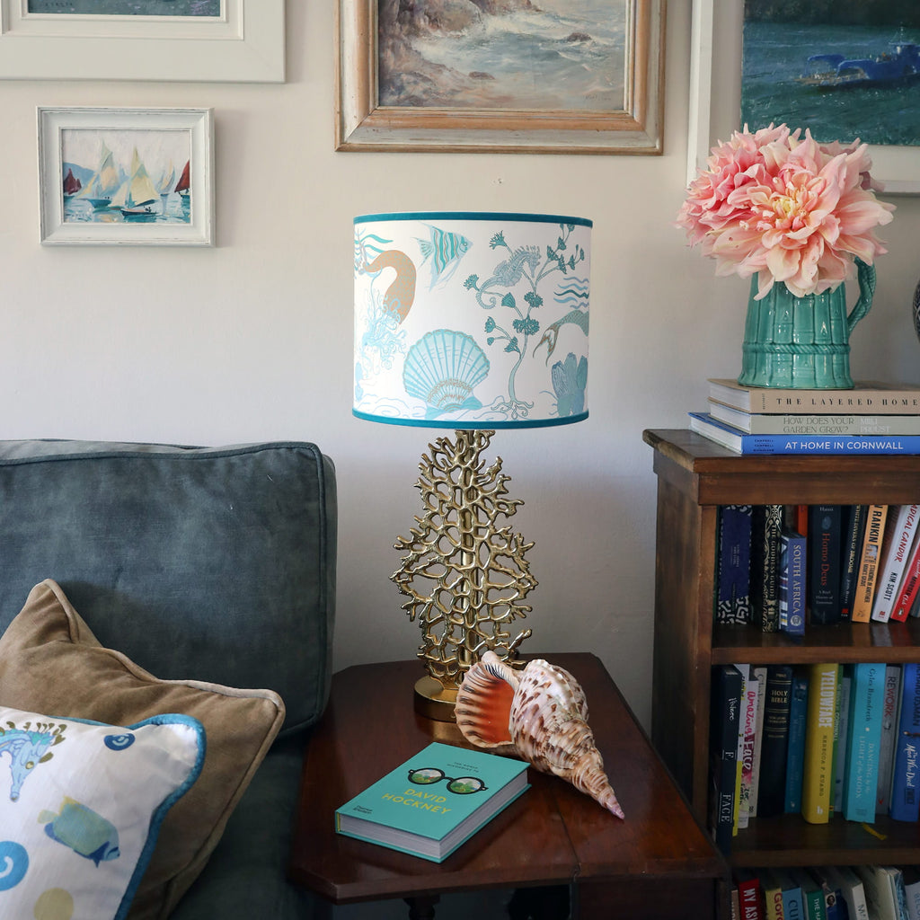Cream Cornwall Mermaid lampshade on a gold coral lamp base, styled with coastal artwork, fresh flowers and seaside accessories in a relaxed coastal living room.