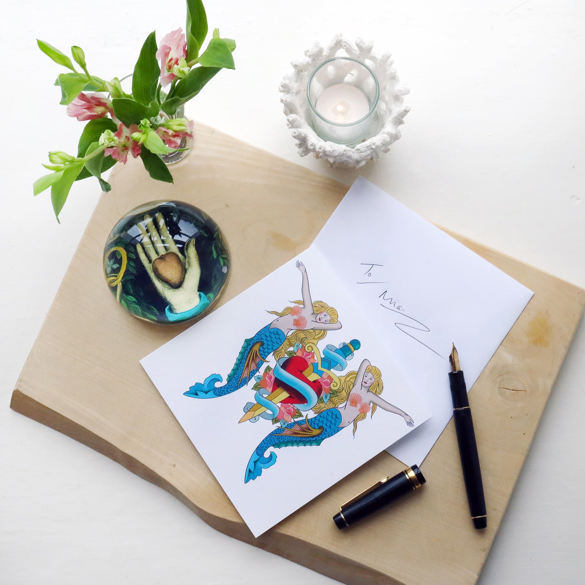White greeting card with illustrated mermaids & daggers tattoo inspired design on, lying on a wooden block with fountain pen, paperweight and flowers and hand written envelope.