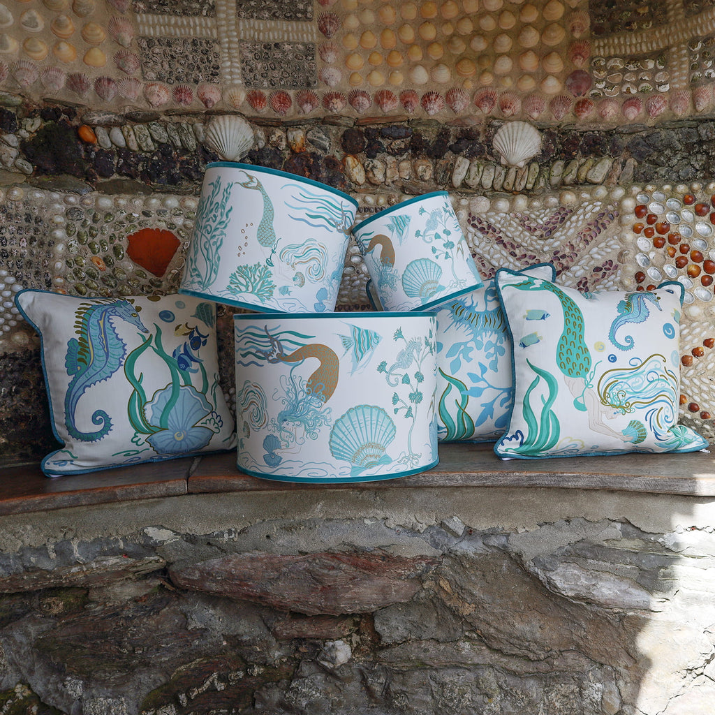 Cream Cornwall Mermaid Collection featuring coordinating cushions and lampshades, styled on a rustic stone bench against a shell-covered wall inspired by the Cornish coast.