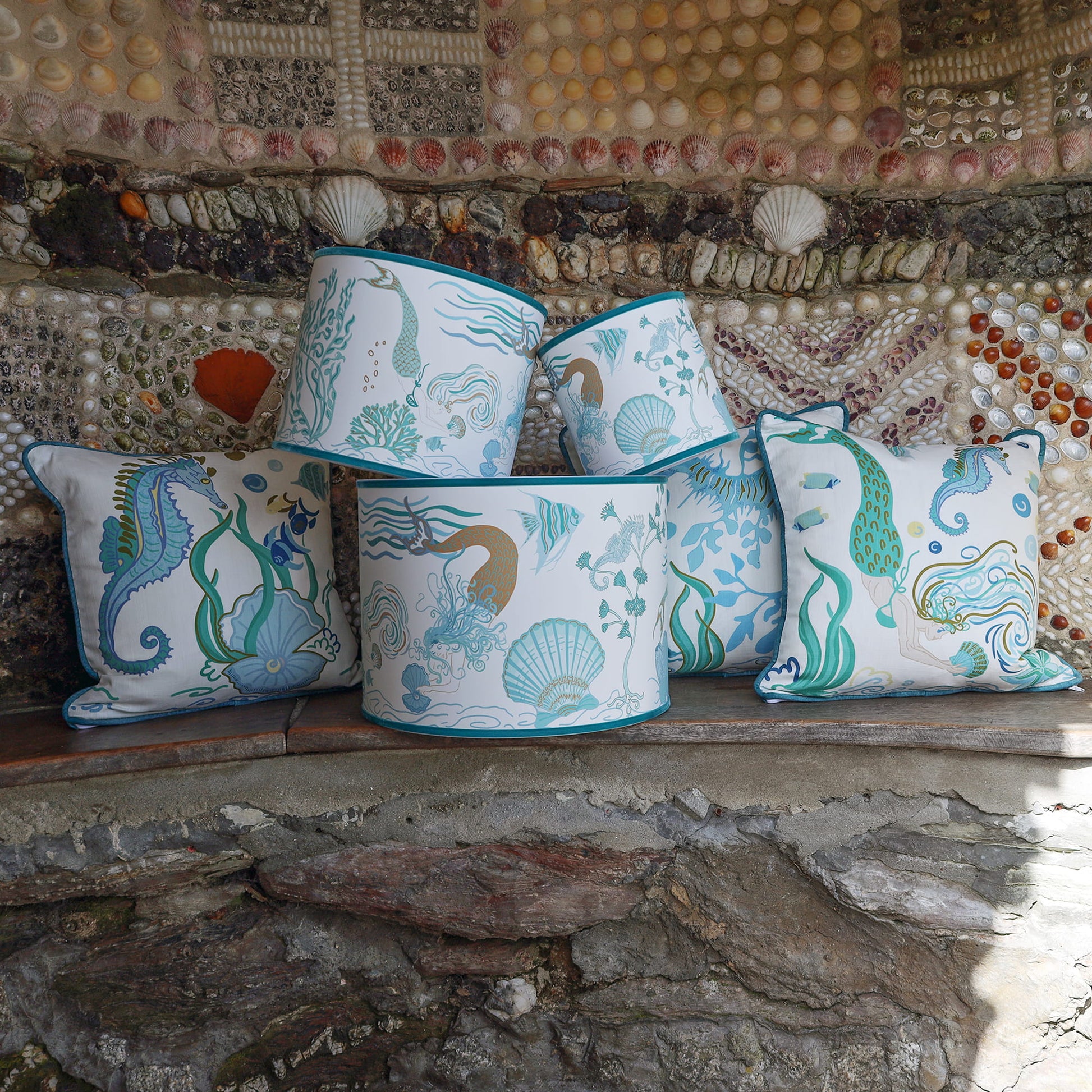 Cream Cornwall Mermaid Collection featuring coordinating cushions and lampshades, styled on a rustic stone bench against a shell-covered wall inspired by the Cornish coast.