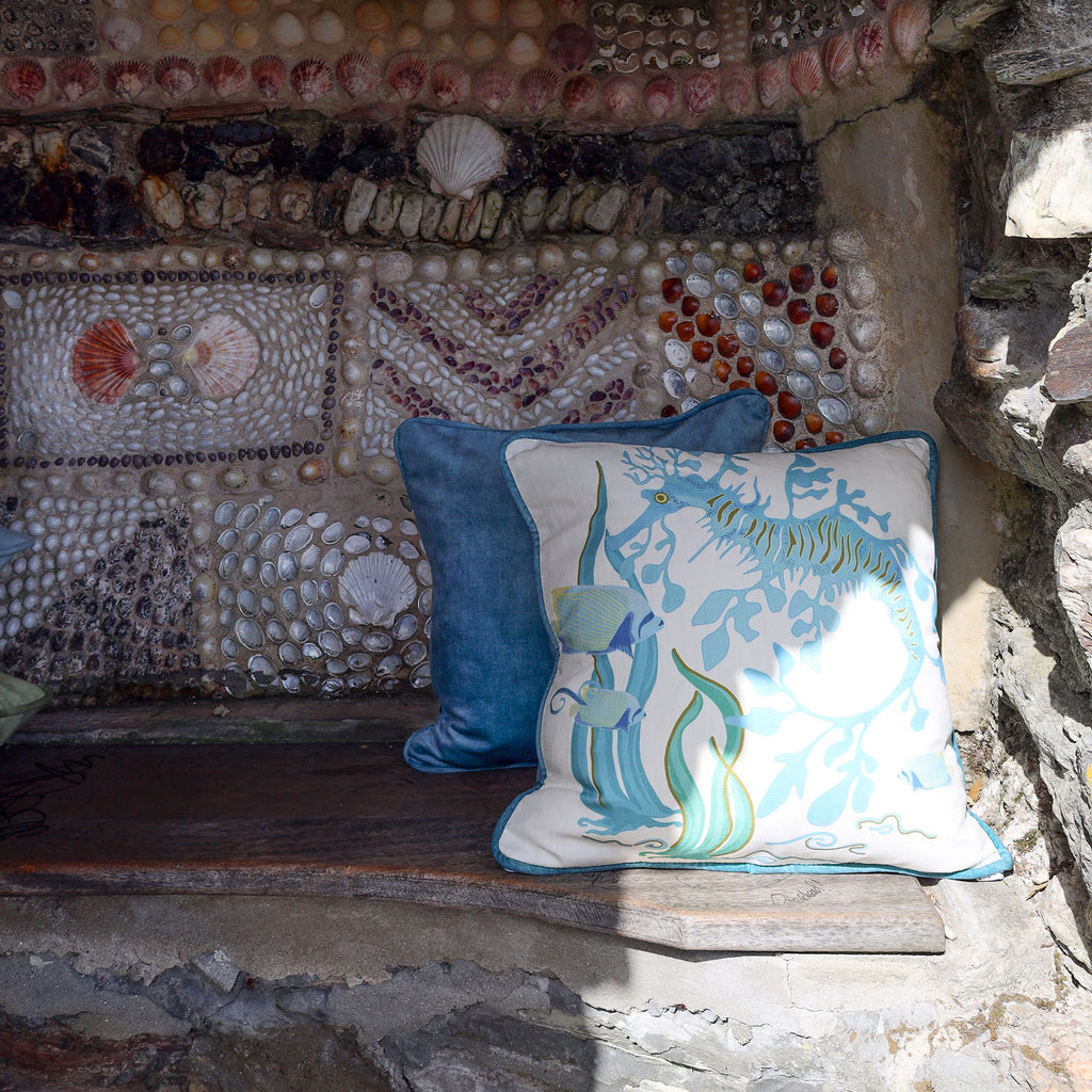 Cream Cornwall Sea Horse cushion paired with a deep blue velvet cushion, styled on a rustic stone bench against a shell-covered wall for a timeless coastal-inspired interior.