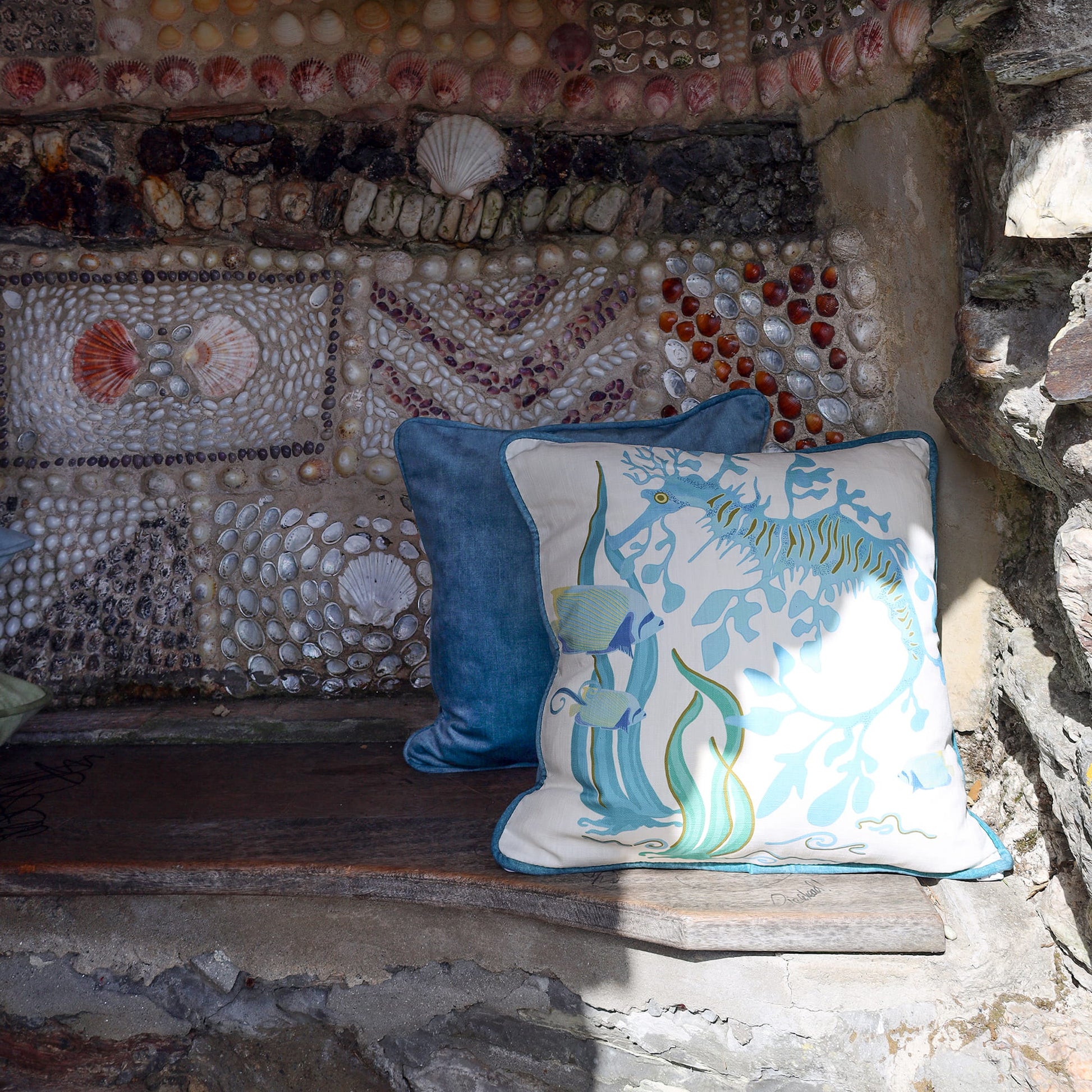 Cream Cornwall Sea Horse cushion paired with a deep blue velvet cushion, styled on a rustic stone bench against a shell-covered wall for a timeless coastal-inspired interior.