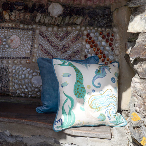 Cream Cornwall Mermaid cushion paired with a deep blue velvet cushion, styled on a rustic stone bench against a shell-covered wall for a relaxed coastal look.