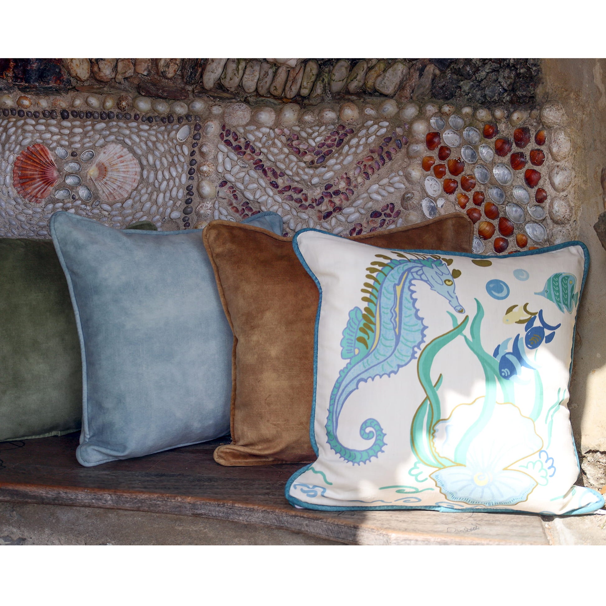 Cream Cornwall Seahorse cushion paired with velvet cushions in sage, sky blue and sand, styled on a rustic stone bench against a shell-covered wall.