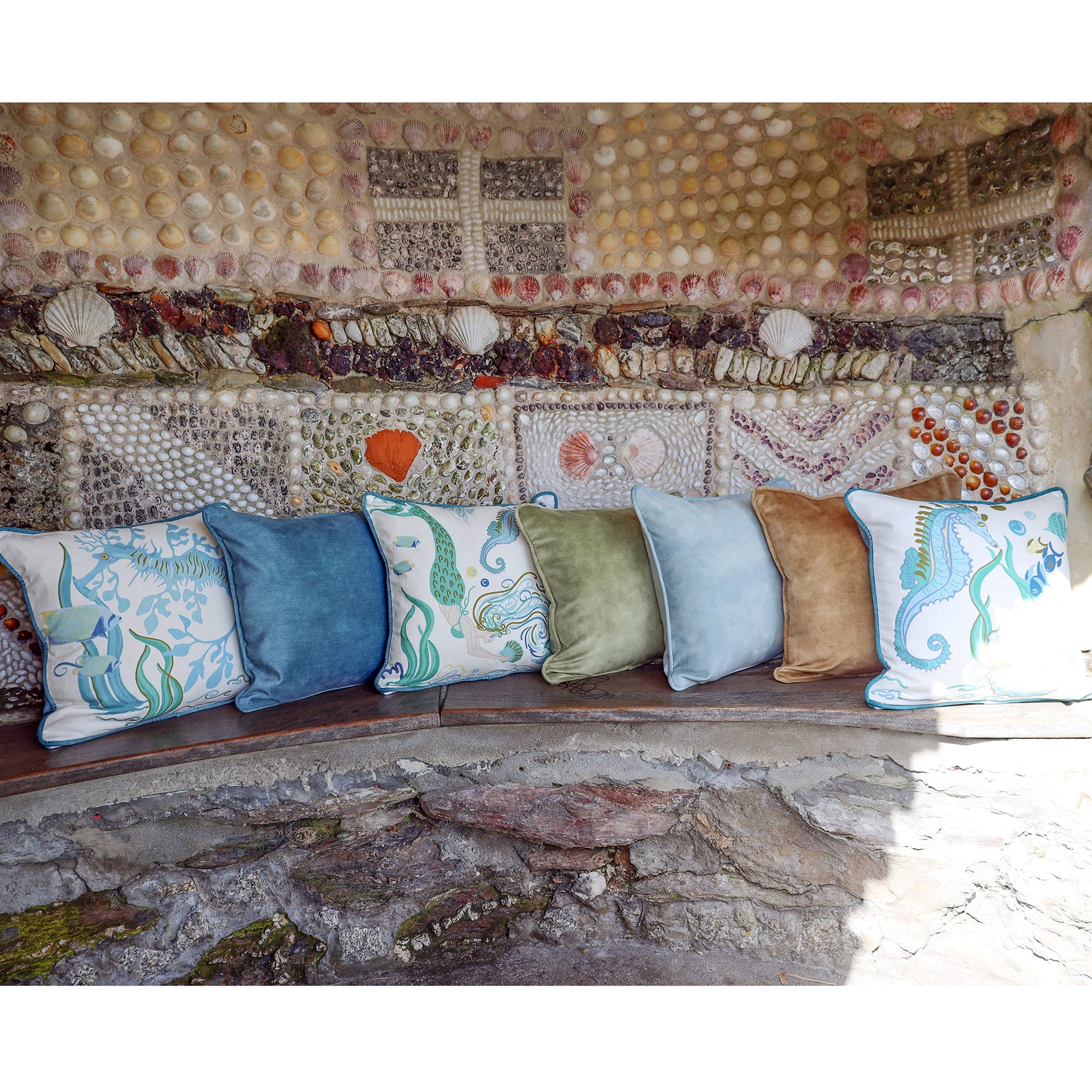 Cream Cornwall coastal cushions in soft blues, greens and sand tones, styled on a rustic stone bench against a shell-covered wall inspired by the Cornish coast.