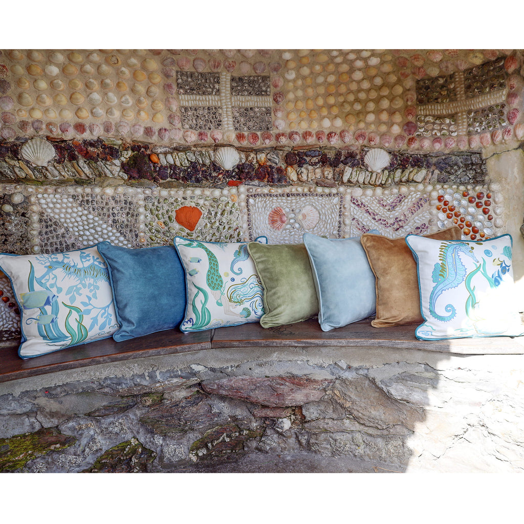 Cream Cornwall coastal cushions in soft blues, greens and sand tones, styled on a rustic stone bench against a shell-covered wall inspired by the Cornish coast.