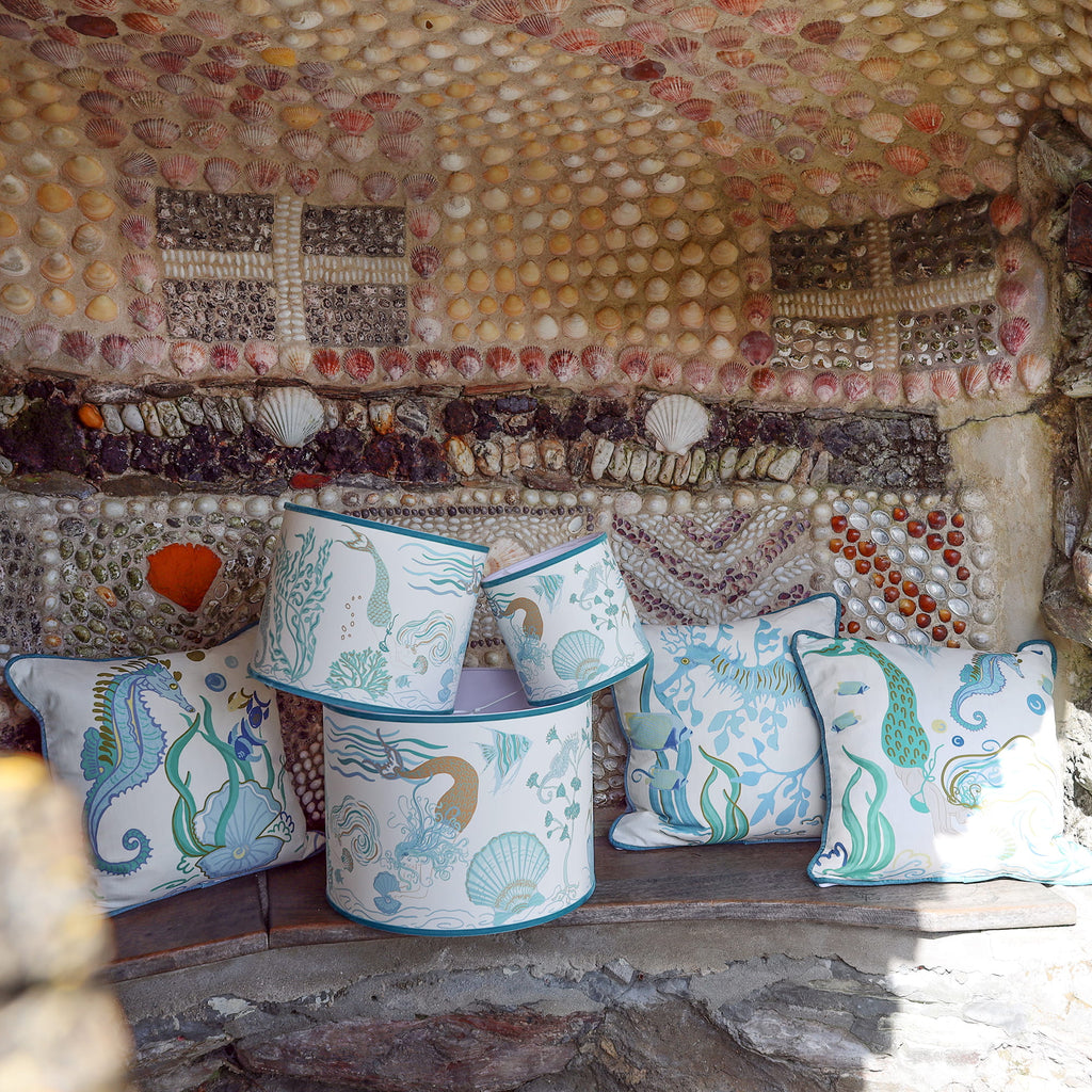 Cream Cornwall Mermaid Collection featuring coordinating cushions and lampshades, styled on a rustic stone bench against a shell-covered wall inspired by the Cornish coast.