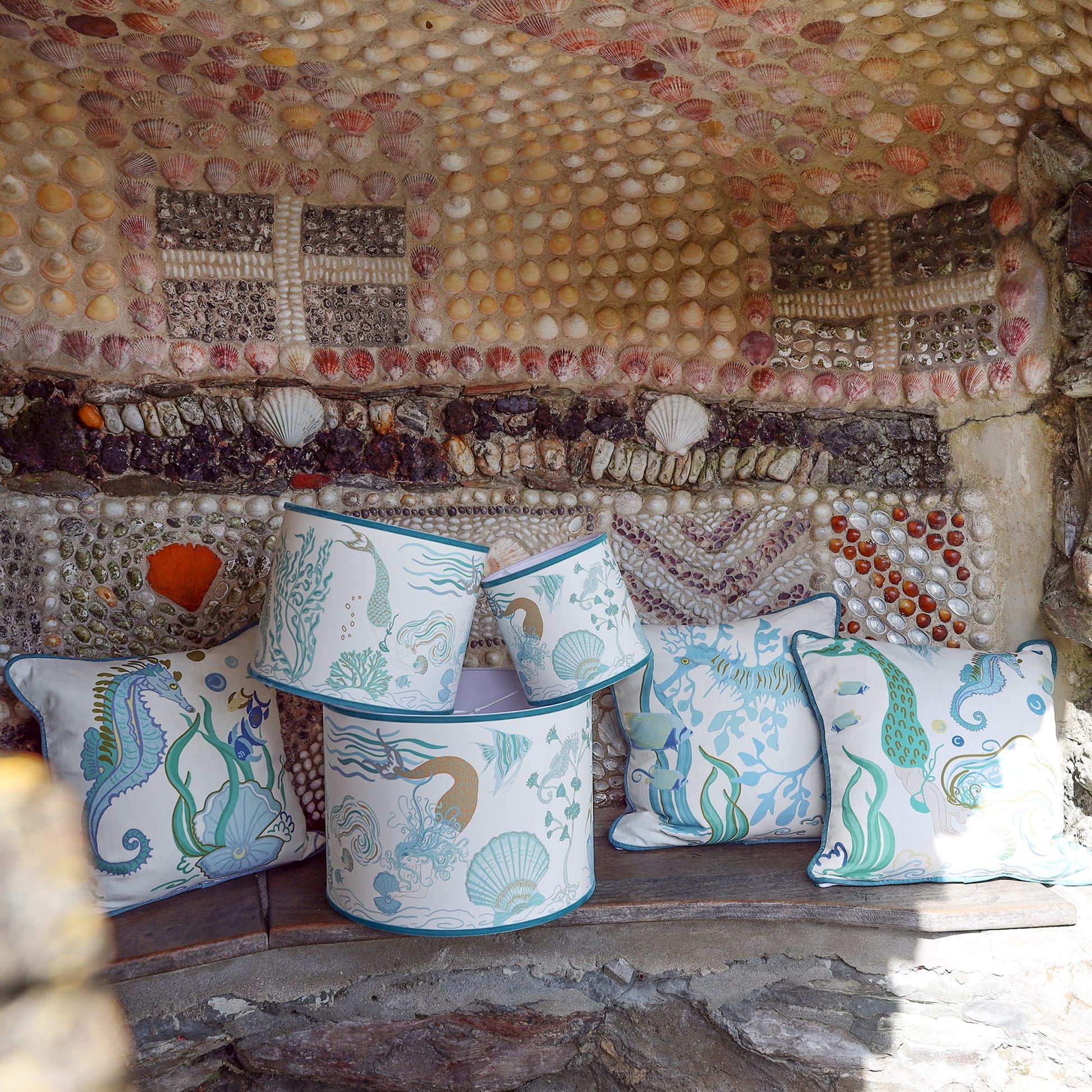 Cream Cornwall Mermaid Collection featuring coordinating cushions and lampshades, styled on a rustic stone bench against a shell-covered wall inspired by the Cornish coast.