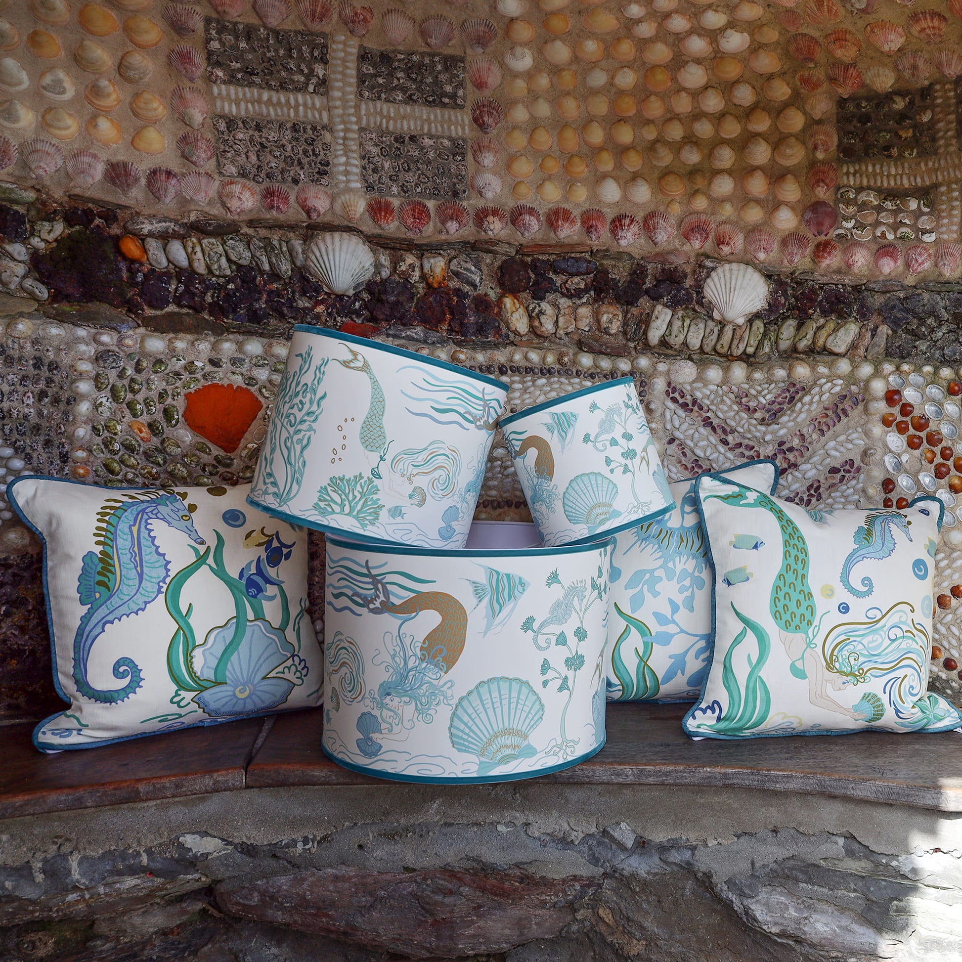 Cream Cornwall Mermaid Collection featuring coordinating cushions and lampshades, styled on a rustic stone bench against a shell-covered wall inspired by the Cornish coast.