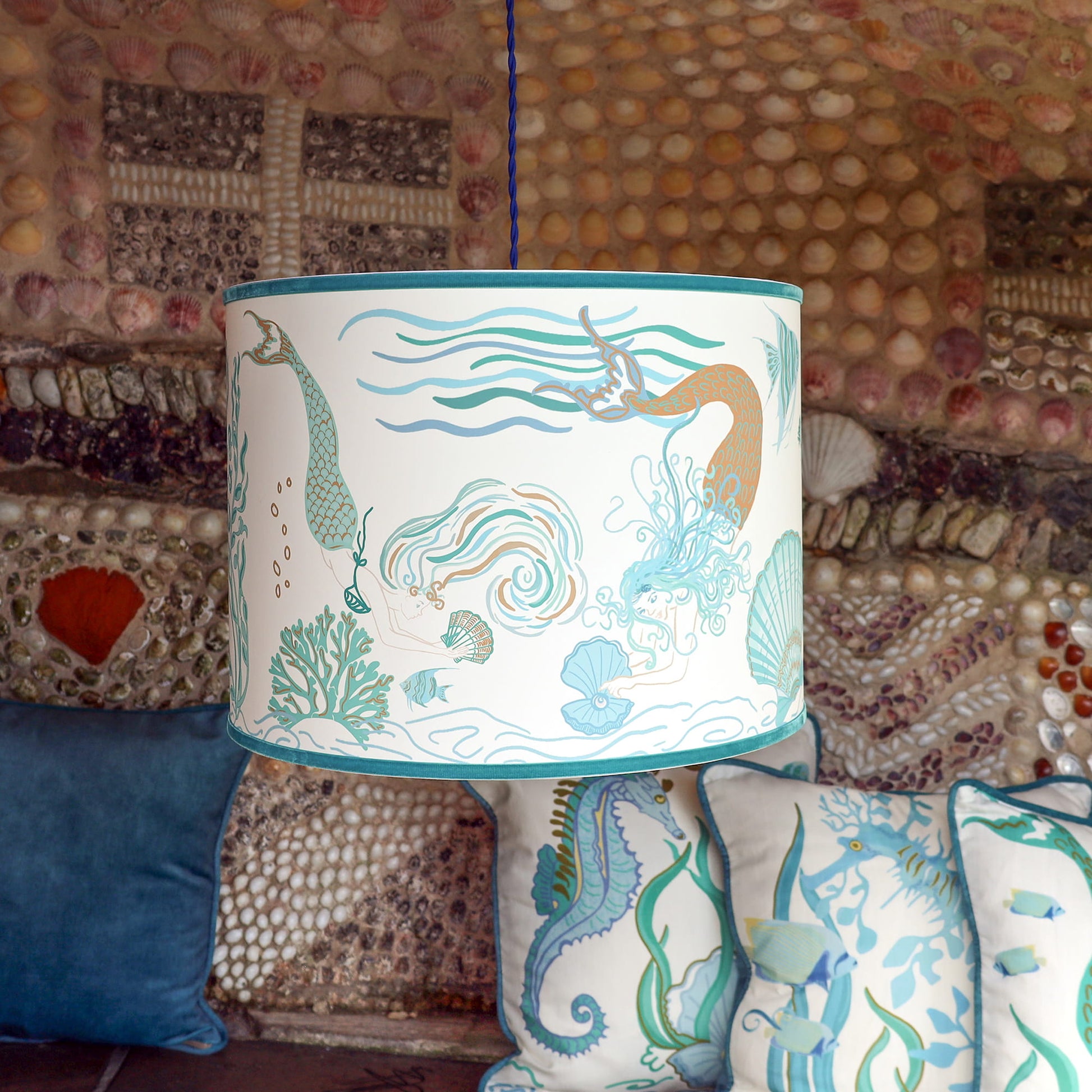 Cream Cornwall Mermaid lampshade hanging above coordinating coastal cushions, featuring an elegant mermaid design in soft blue, turquoise and sand tones against a shell-covered wall.