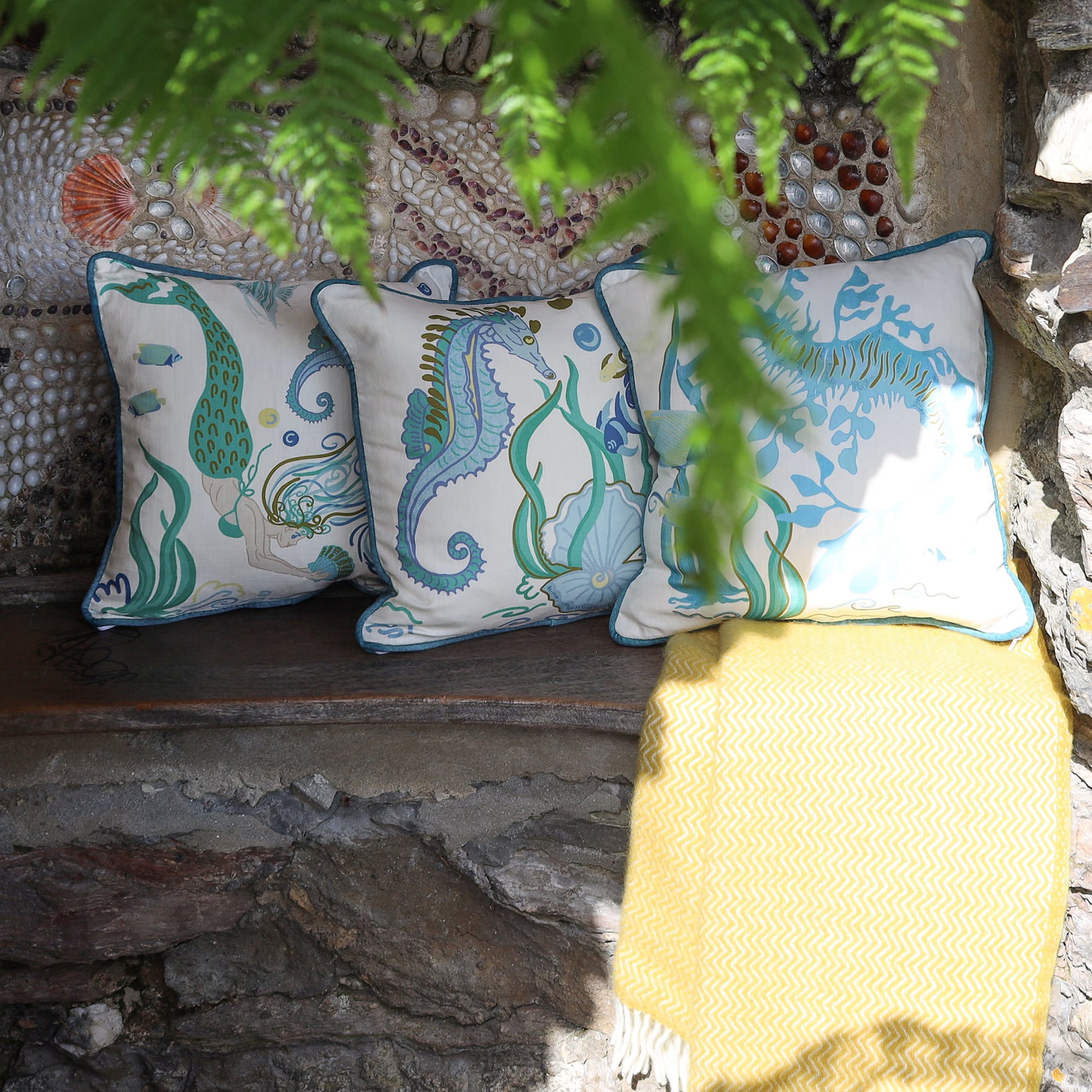 Cream Cornwall Mermaid, Seahorse and Sea Horse cushions styled with a soft yellow throw on a rustic stone bench, creating a relaxed coastal setting beneath trailing ferns.