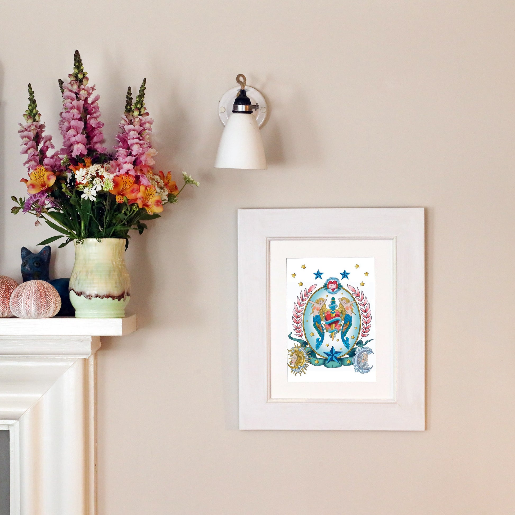 A framed nautical-themed illustration featuring mermaids, stars, and maritime elements, hanging on a pale wall above a mantel. The mantel is decorated with a vase of colorful flowers, ceramic ornaments, and a cat figurine. A white wall-mounted lamp is positioned above the frame.