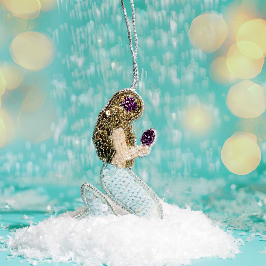 Mermaid ornament with glittery hair and blue tail on a snowy base against a blurred light background