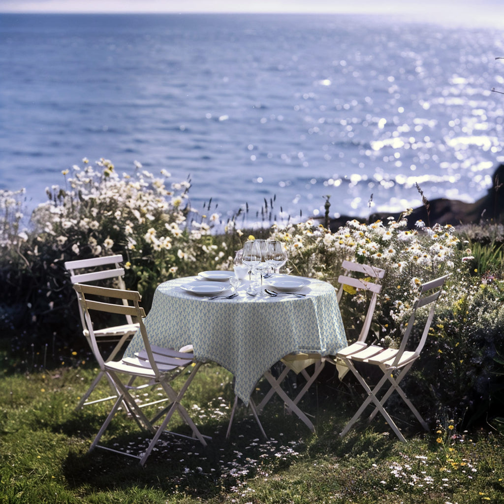 An idyllic outdoor dining setup on a grassy seaside garden, overlooking a sparkling ocean. A round table is covered with a soft blue and white patterned tablecloth, surrounded by four white folding chairs. The table is set with elegant white plates, glasses, and cutlery. Daisies and wildflowers bloom around the scene, adding a natural and romantic charm to the coastal atmosphere.