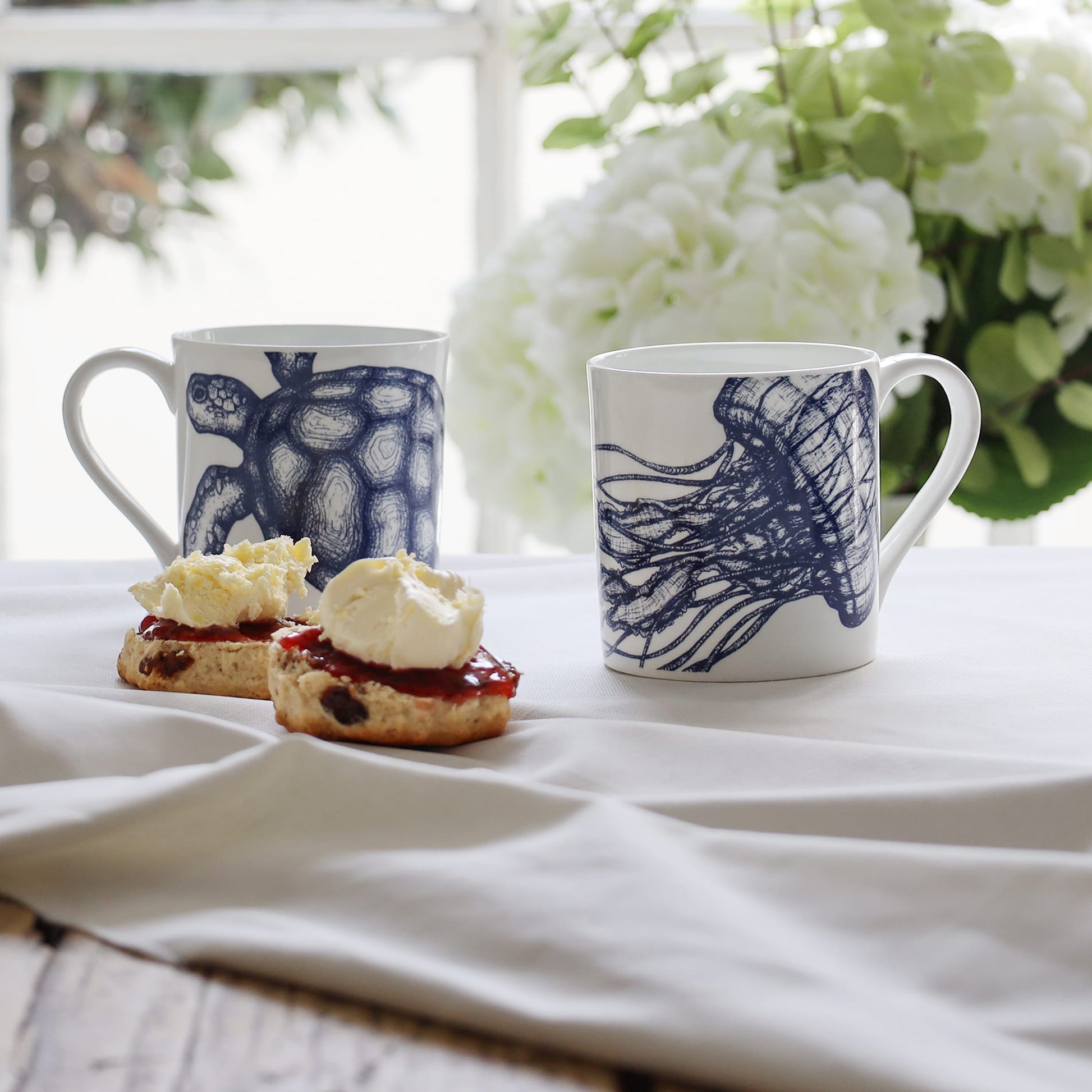 Two mugs with blue jellyfish & turtle designs on a white tablecloth with scones.