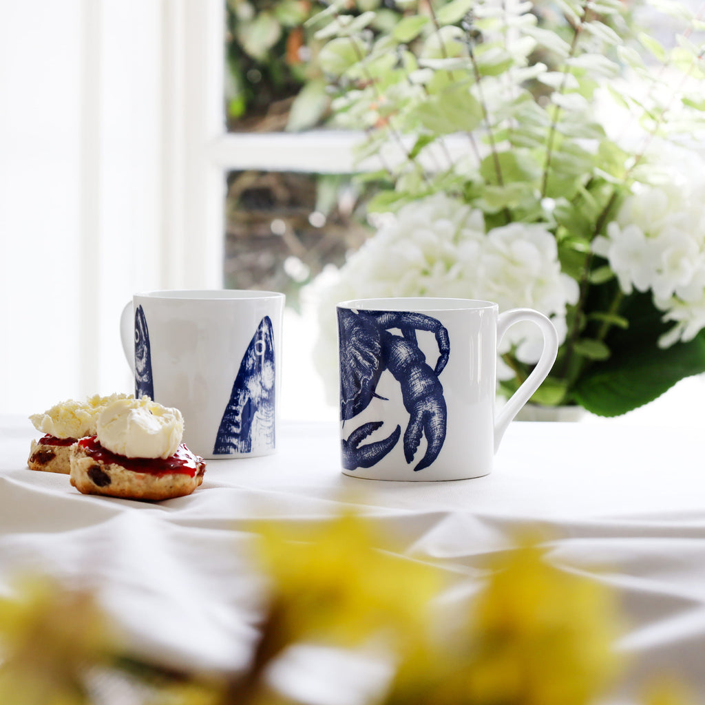 Two mugs with crab & mackerel heads design on a table with scones and jam, flowers in the background