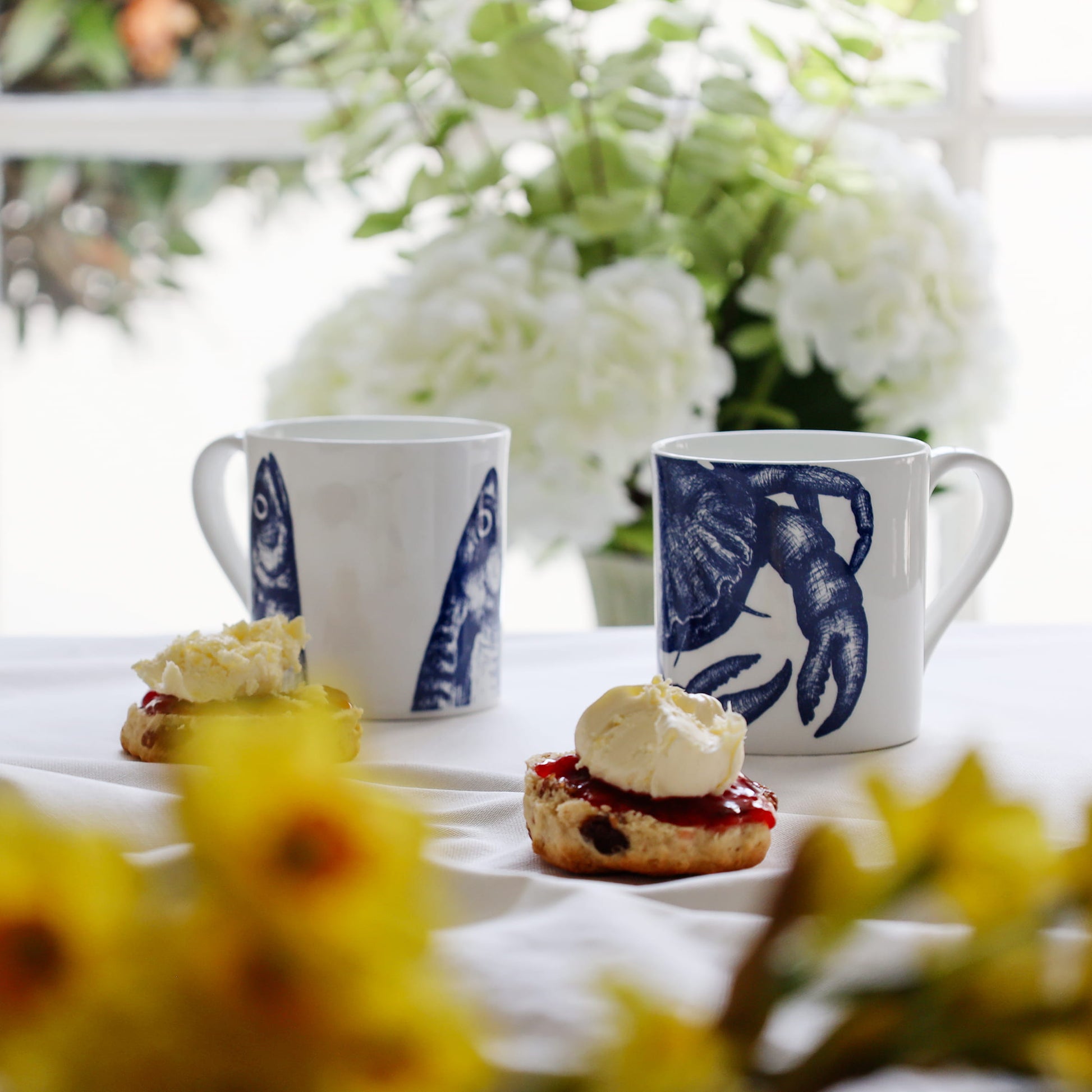 Two mugs with blue crab & mackerel heads design on a table with scones and cream, surrounded by flowers.