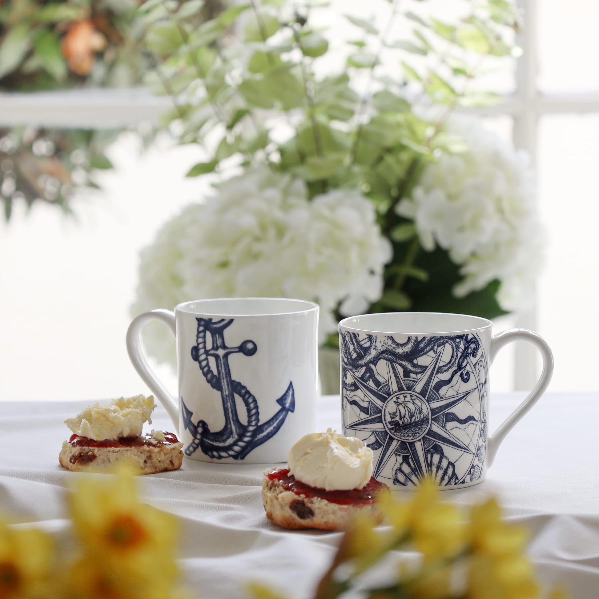 Two mugs with nautical designs on a table with scones and flowers.