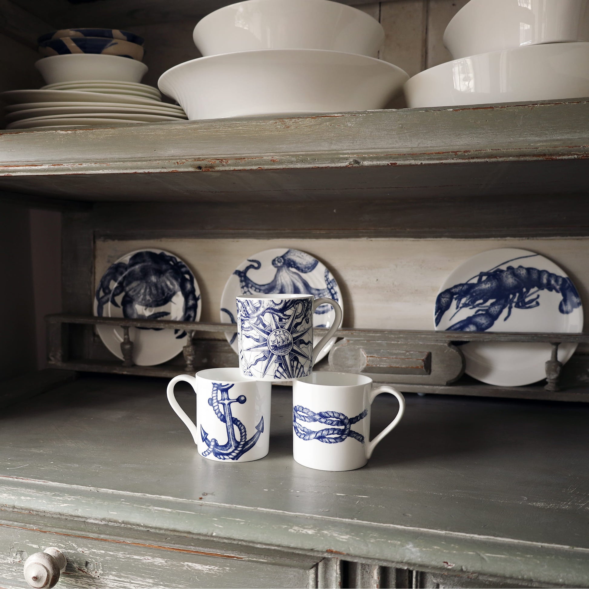 White mugs with blue designs on a wooden shelf with ceramic plates in the background.